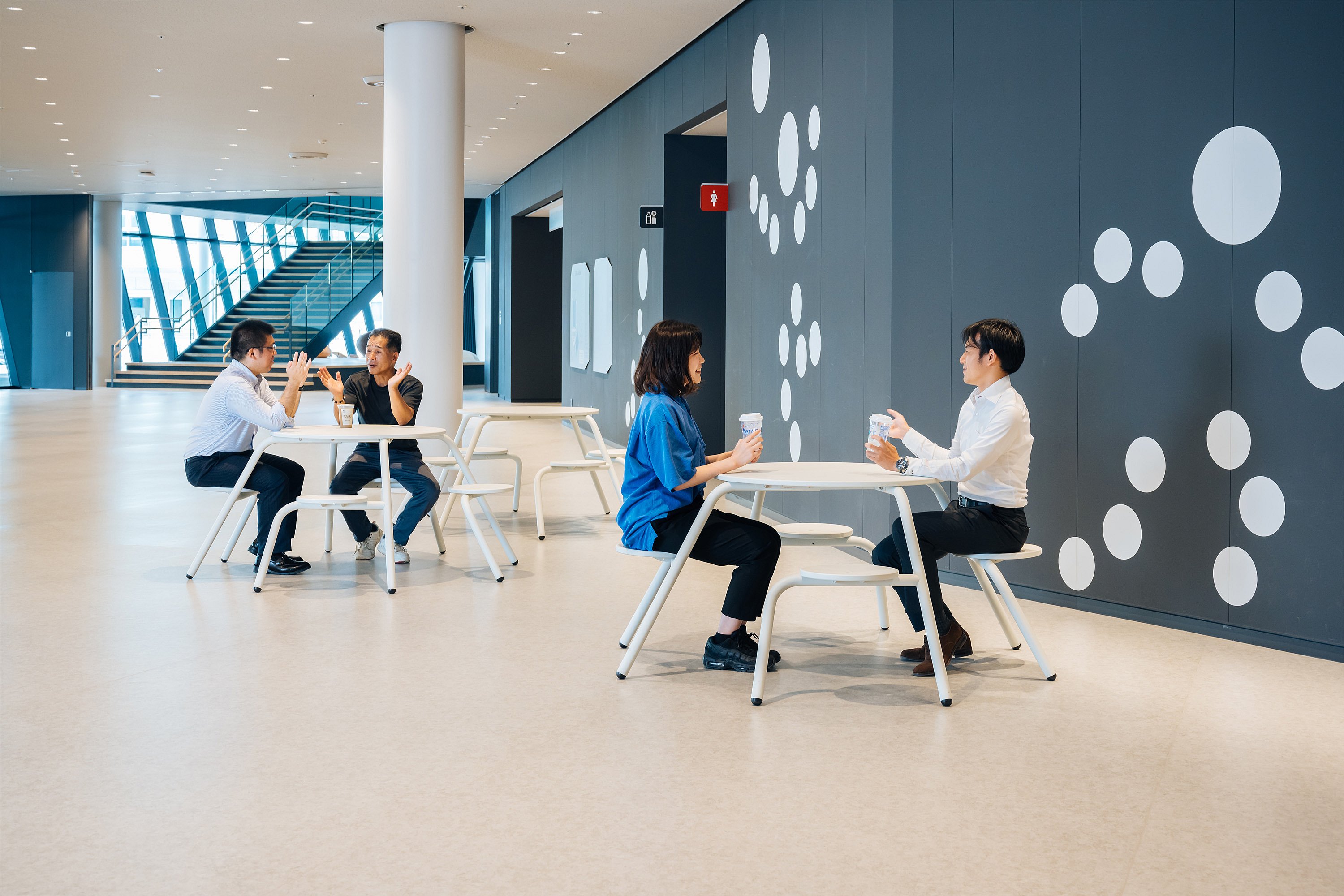 Spacious indoor concourse with two pairs of people seated at white round virus tables with integrated stools, holding takeaway cups beside a dark wall decorated with large white dots.