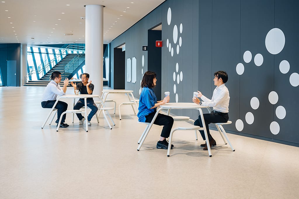 Spacious indoor concourse with two pairs of people seated at white round virus tables with integrated stools, holding takeaway cups beside a dark wall decorated with large white dots.