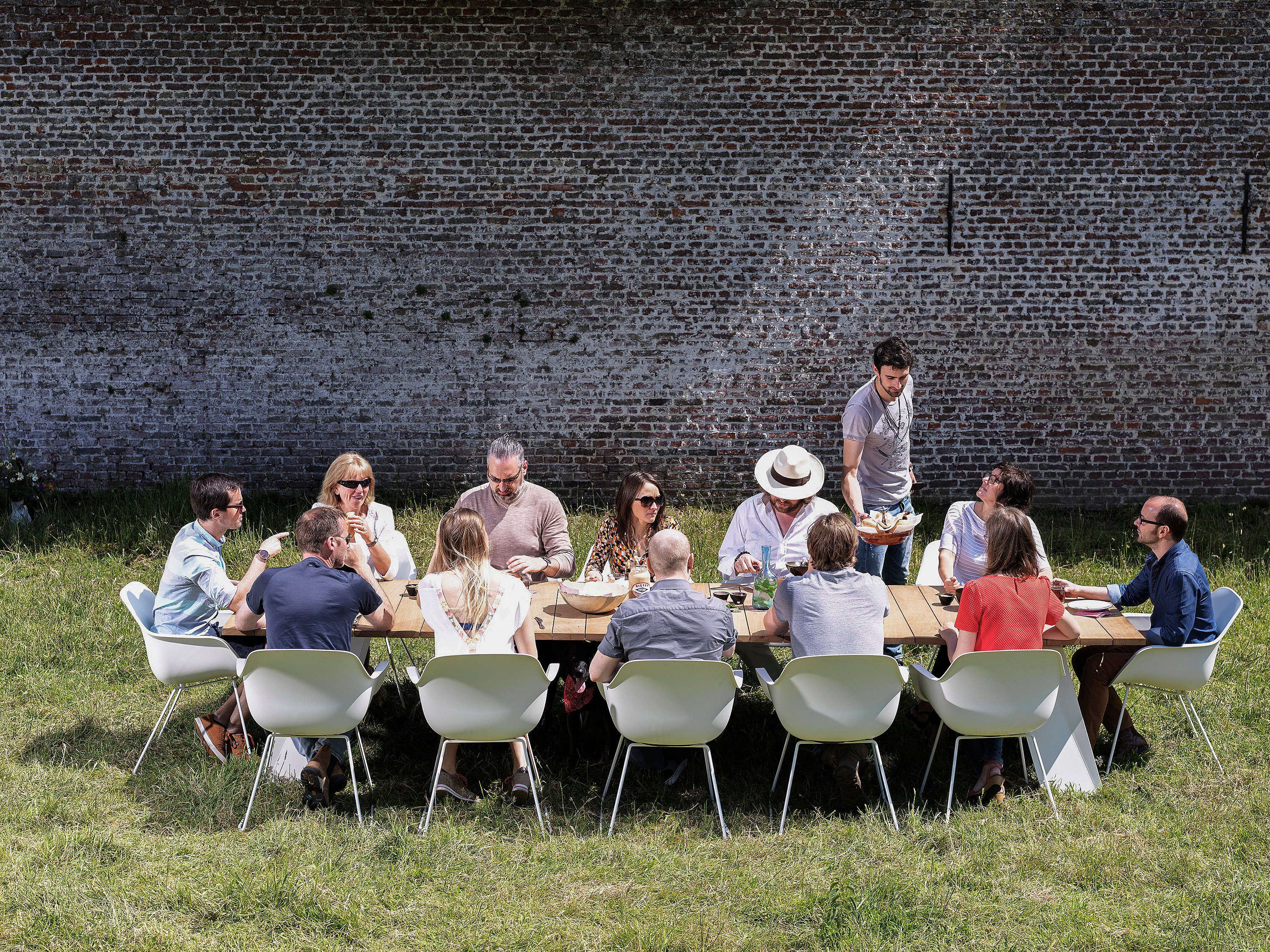 A group of colleagues share lunch around a long wooden Pontsūn table with white Captain’s Chairs in a sunny walled garden.