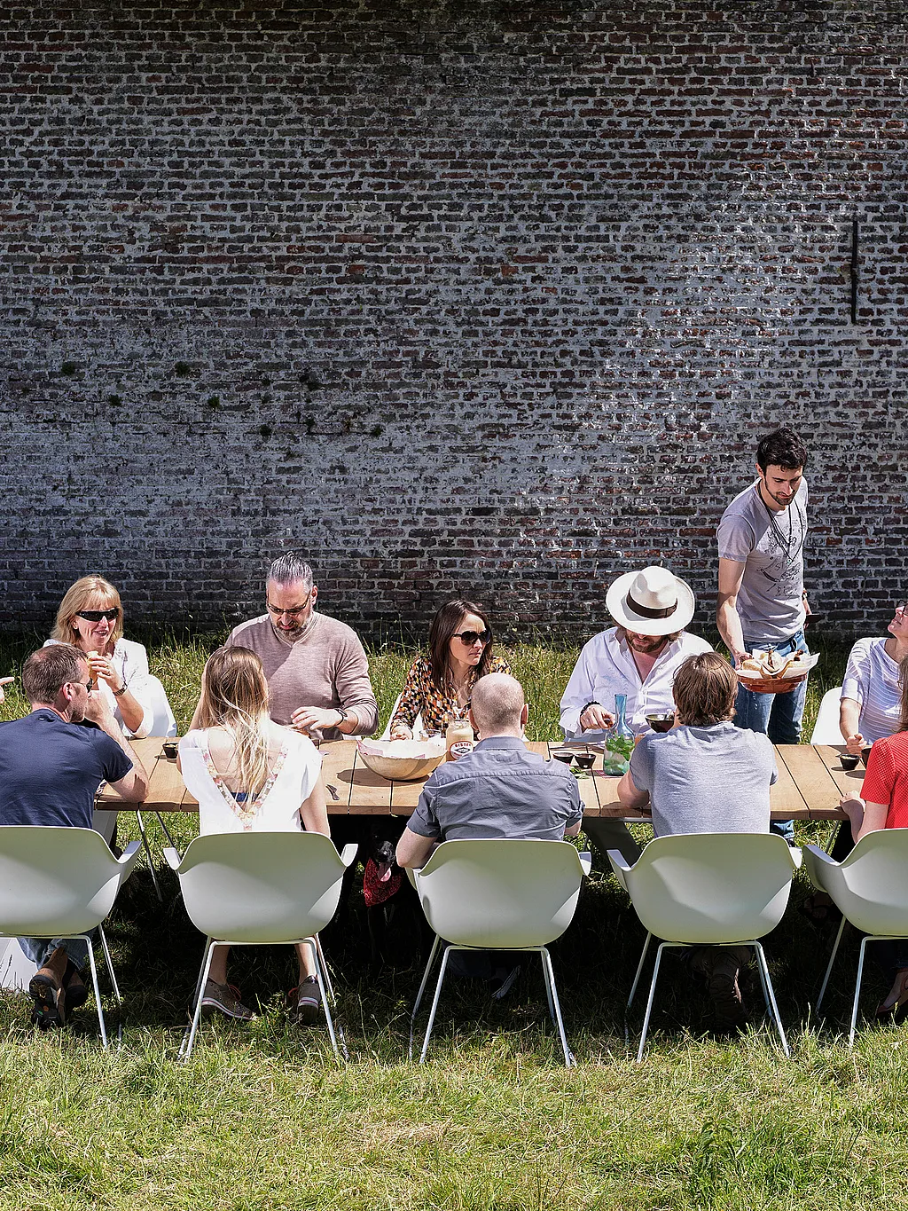 A group of colleagues share lunch around a long wooden Pontsūn table with white Captain’s Chairs in a sunny walled garden.