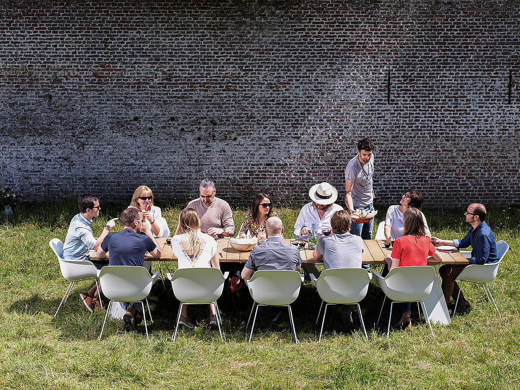 Eine Gruppe von Kollegen teilt ihr Mittagessen an einem langen hölzernen Pontsūn-Tisch mit weißen Kapitänsstühlen in einem sonnigen, ummauerten Garten.