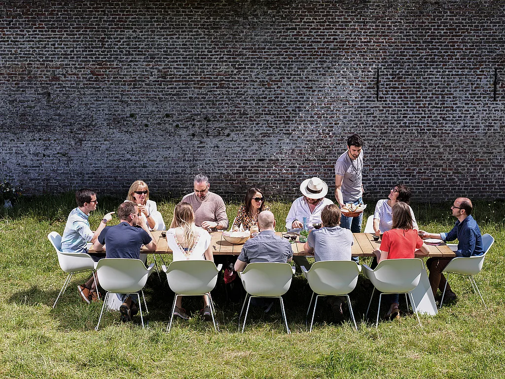 Eine Gruppe von Kollegen teilt ihr Mittagessen an einem langen hölzernen Pontsūn-Tisch mit weißen Kapitänsstühlen in einem sonnigen, ummauerten Garten.