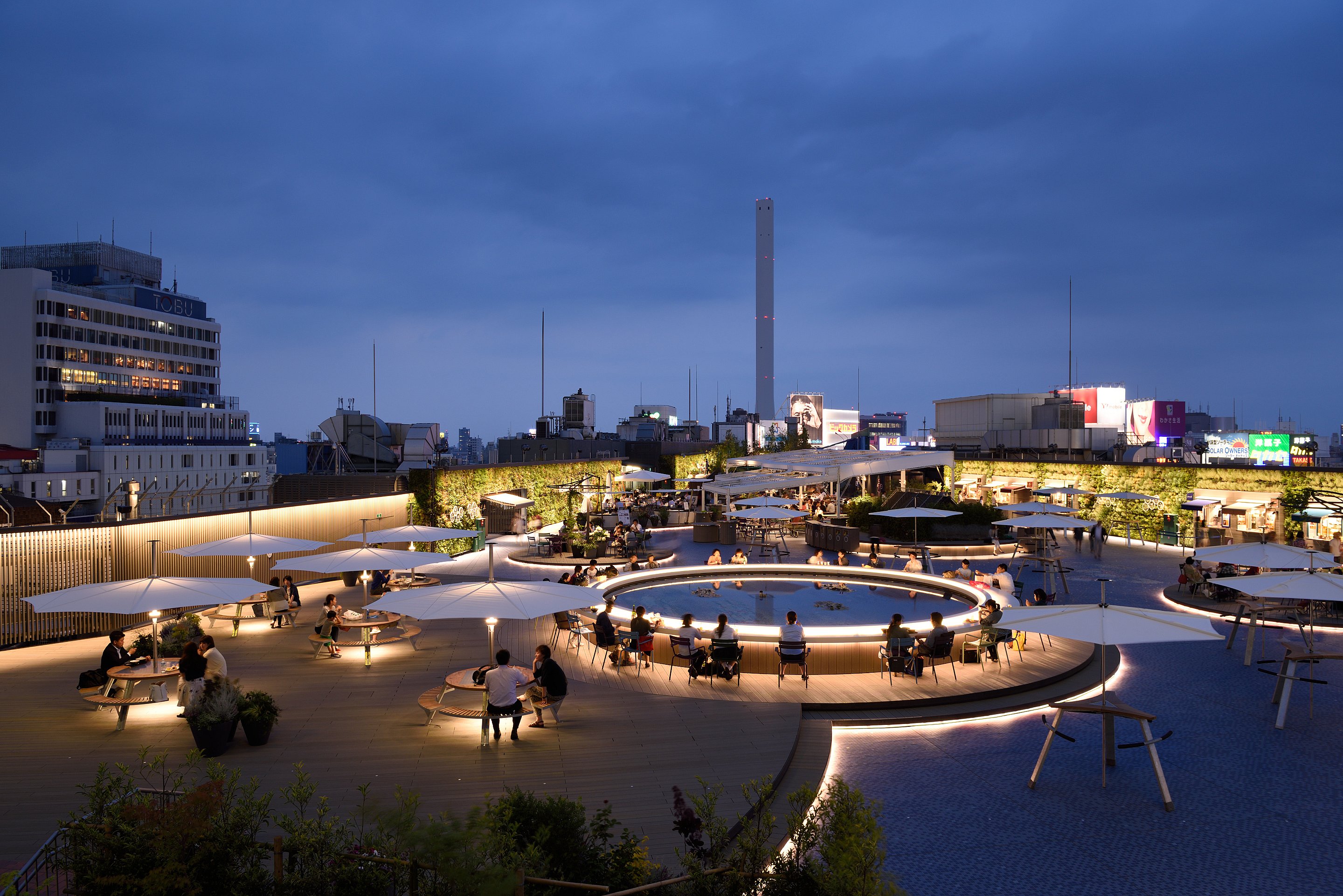 In der Abenddämmerung sitzen Besucher an beleuchteten Gargantua-Picknicktischen unter großen weißen Sonnenschirmen auf der Dachterrasse, im Hintergrund leuchten Gebäude aus Tokio.