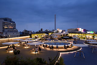 In der Abenddämmerung sitzen Besucher an beleuchteten Gargantua-Picknicktischen unter großen weißen Sonnenschirmen auf der Dachterrasse, im Hintergrund leuchten Gebäude aus Tokio.