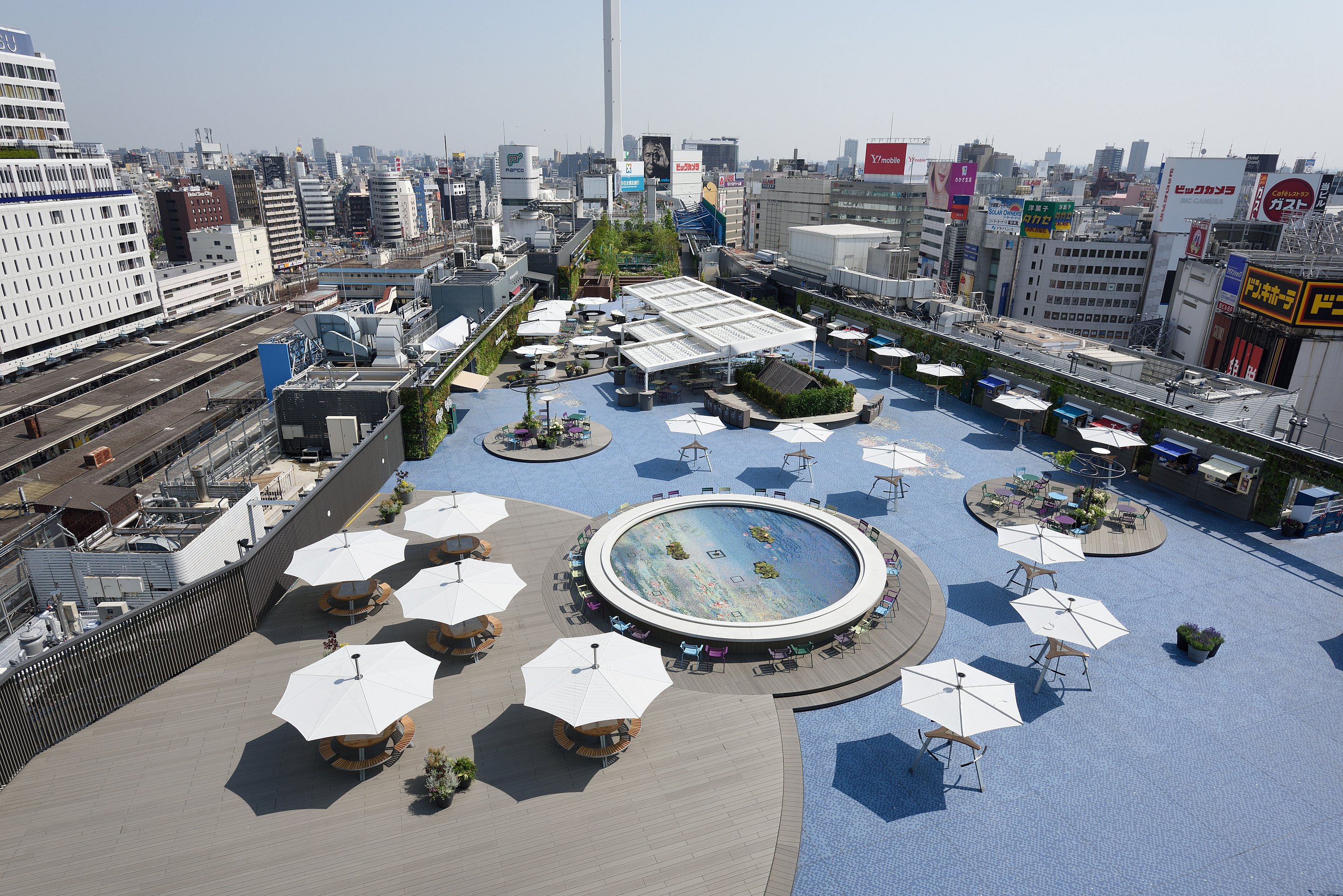 Vue en contre-plongée du jardin sur le toit de Seibu Ikebukuro à Tokyo, avec un pavage en mosaïque bleue, une fontaine ronde, des bordures plantées, des tables partagées et des parasols blancs répartis sur la terrasse.