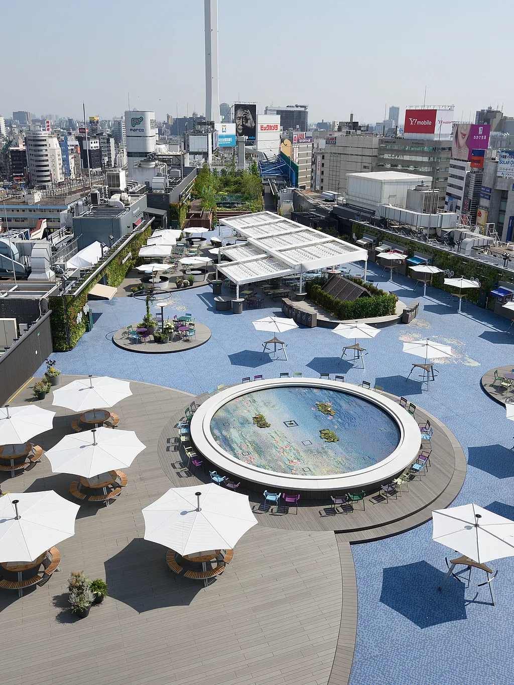 Vue en contre-plongée du jardin sur le toit de Seibu Ikebukuro à Tokyo, avec un pavage en mosaïque bleue, une fontaine ronde, des bordures plantées, des tables partagées et des parasols blancs répartis sur la terrasse.