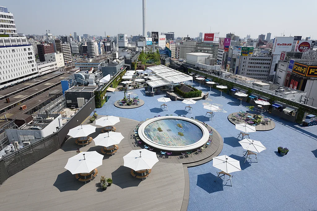 Weitwinkelblick auf den Seibu Ikebukuro-Dachgarten in Tokio mit blauem Mosaikpflaster, einem runden Wasserspiel, bepflanzten Rabatten, gemeinsamen Tischen und weißen Sonnenschirmen, die über die Terrasse verteilt sind.