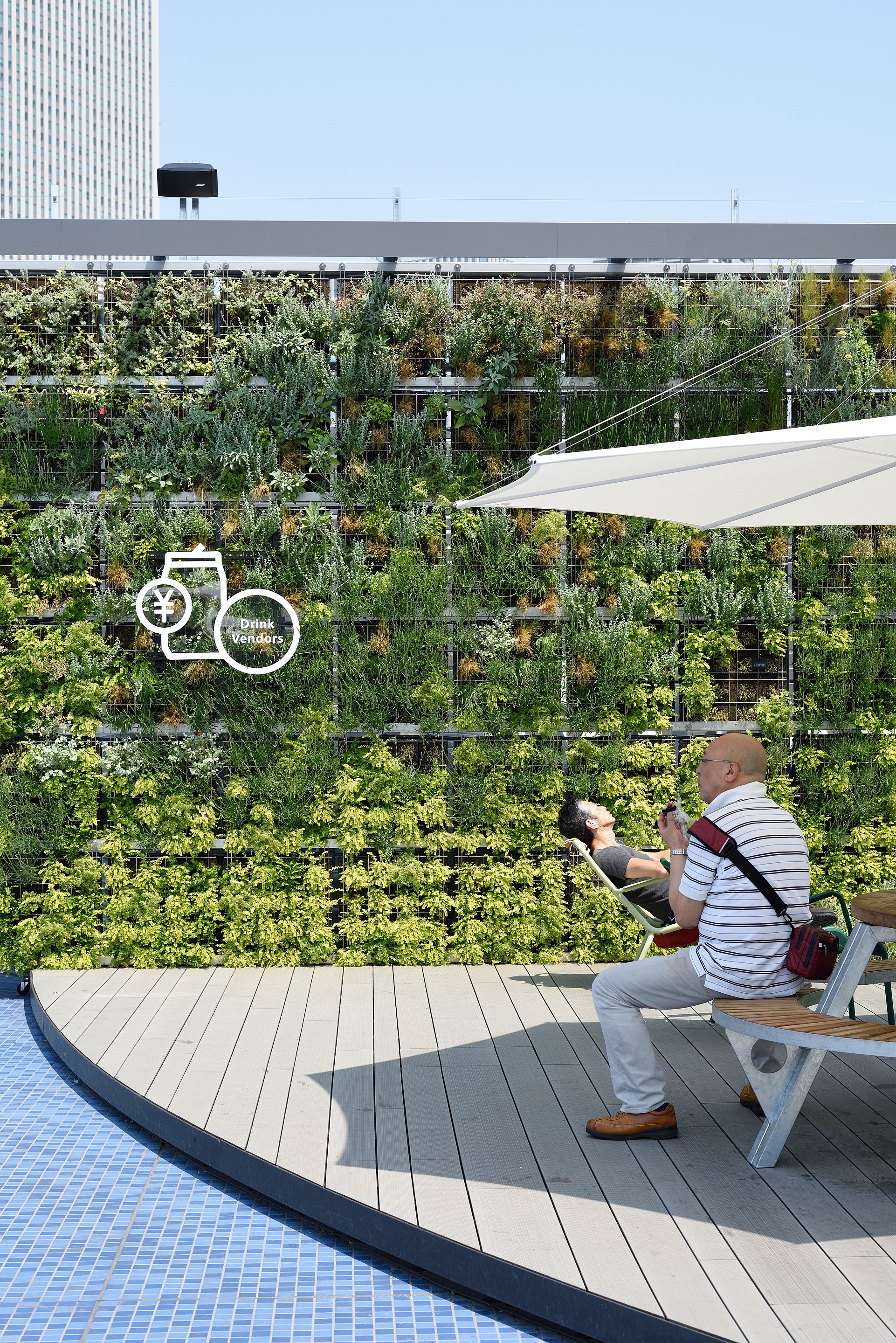 “Ein Besucher sitzt am Rand einer Dachterrasse unter einem weißen Inumbra-Sonnenschirm, während ein anderer neben einem dichten vertikalen Garten liegt.