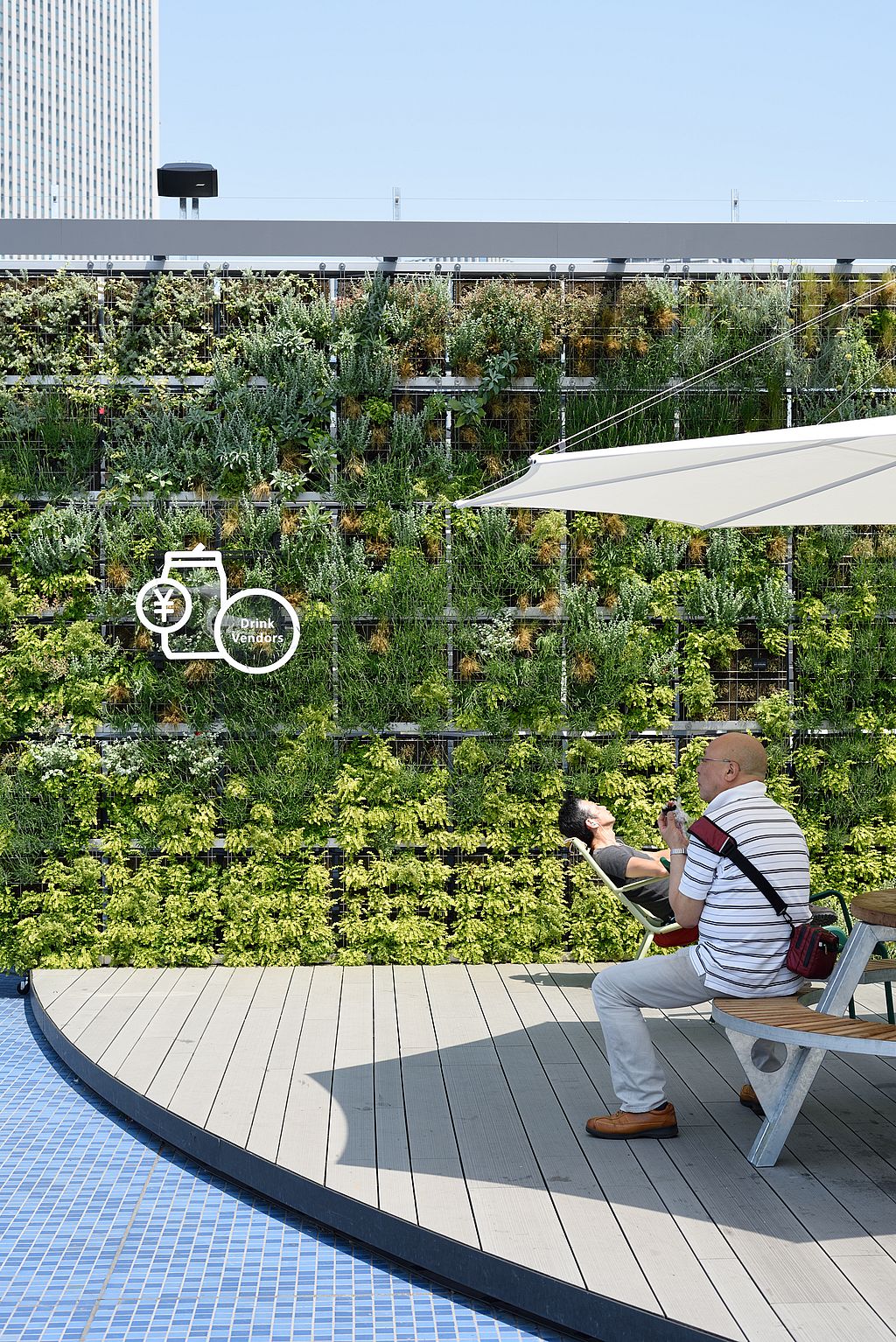 “Ein Besucher sitzt am Rand einer Dachterrasse unter einem weißen Inumbra-Sonnenschirm, während ein anderer neben einem dichten vertikalen Garten liegt.