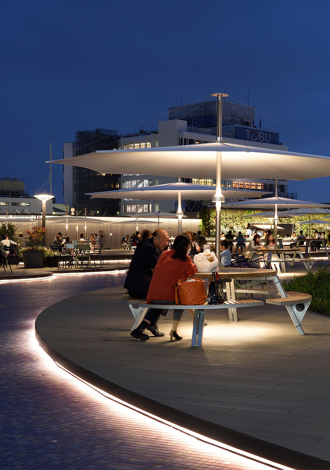 In der Abenddämmerung sitzen Besucher an beleuchteten Gargantua-Picknicktischen unter großen weißen Inumbra-Sonnenschirmen auf der Dachterrasse, im Hintergrund leuchten Gebäude aus Tokio.