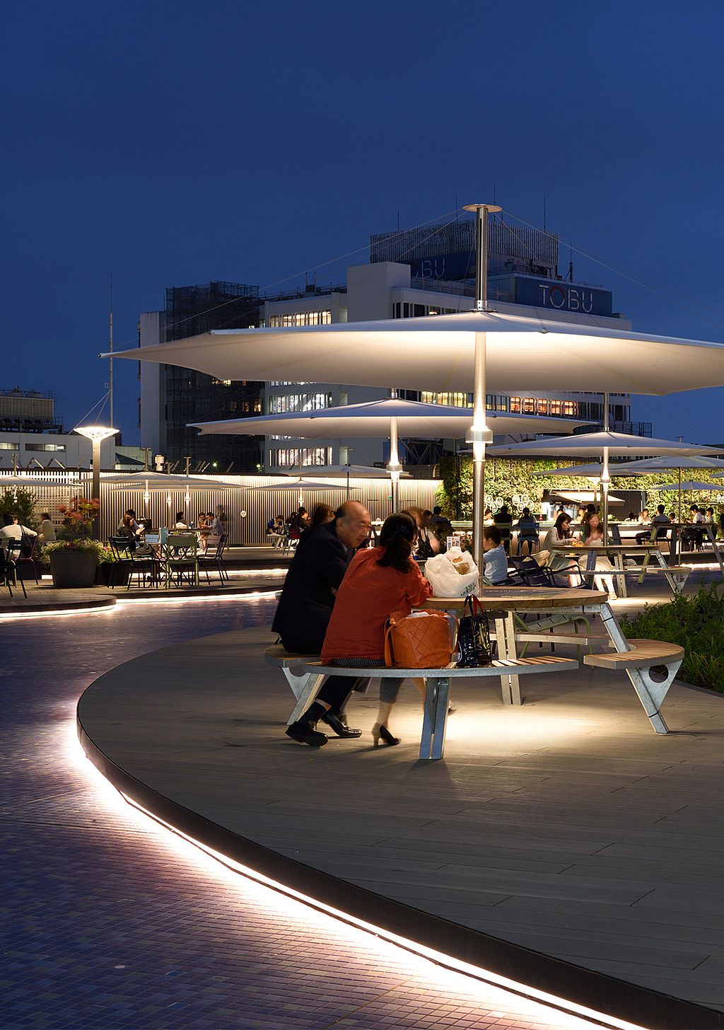 In der Abenddämmerung sitzen Besucher an beleuchteten Gargantua-Picknicktischen unter großen weißen Inumbra-Sonnenschirmen auf der Dachterrasse, im Hintergrund leuchten Gebäude aus Tokio.