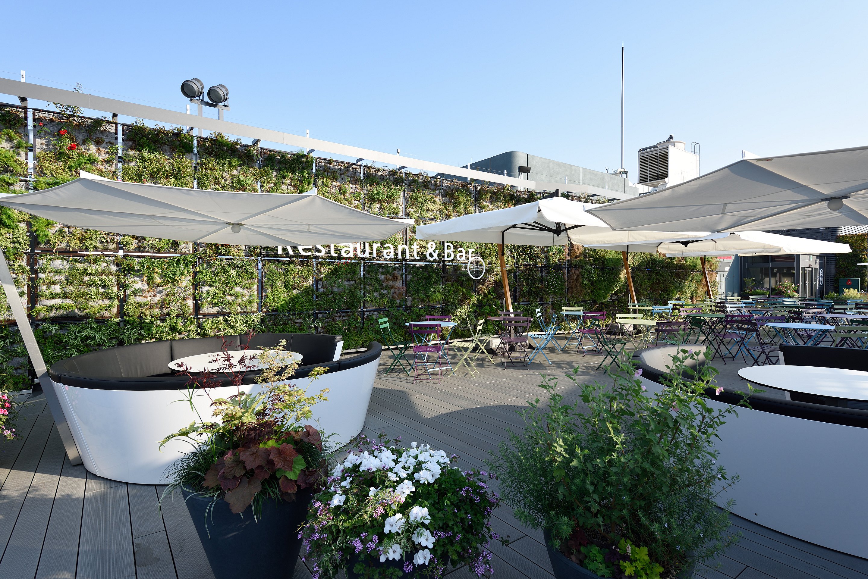 Zwei weiße Kosmos-Sofas stehen unter großen weißen Jalousien auf einer hölzernen Dachterrasse, umgeben von Blumentöpfen, Caféstühlen und einer hohen grünen Wohnwand.