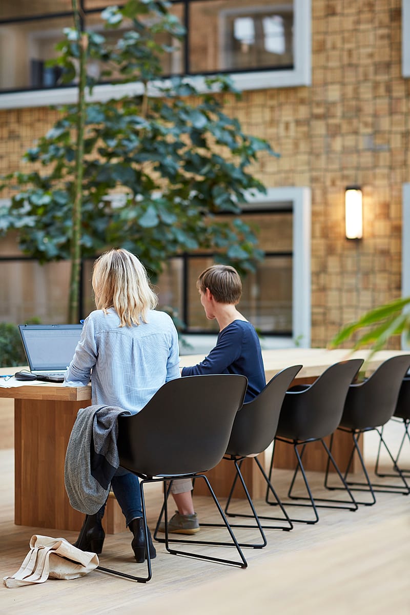 Zwei Personen arbeiten nebeneinander auf schwarzen Captain’s Chairs an einem langen Holztisch in der überdachten Gasse des Gebäudes.“