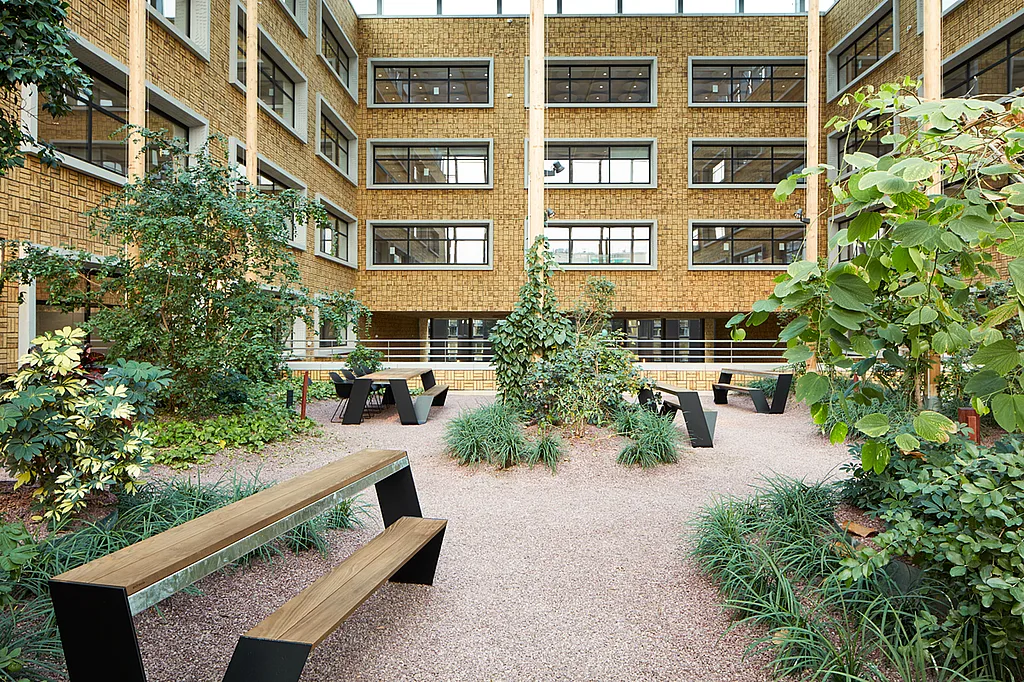 Indoor courtyard of the Herman Teirlinck building with landscaped planting beds, gravel paths and several Hopper benches beneath a high glass roof.
