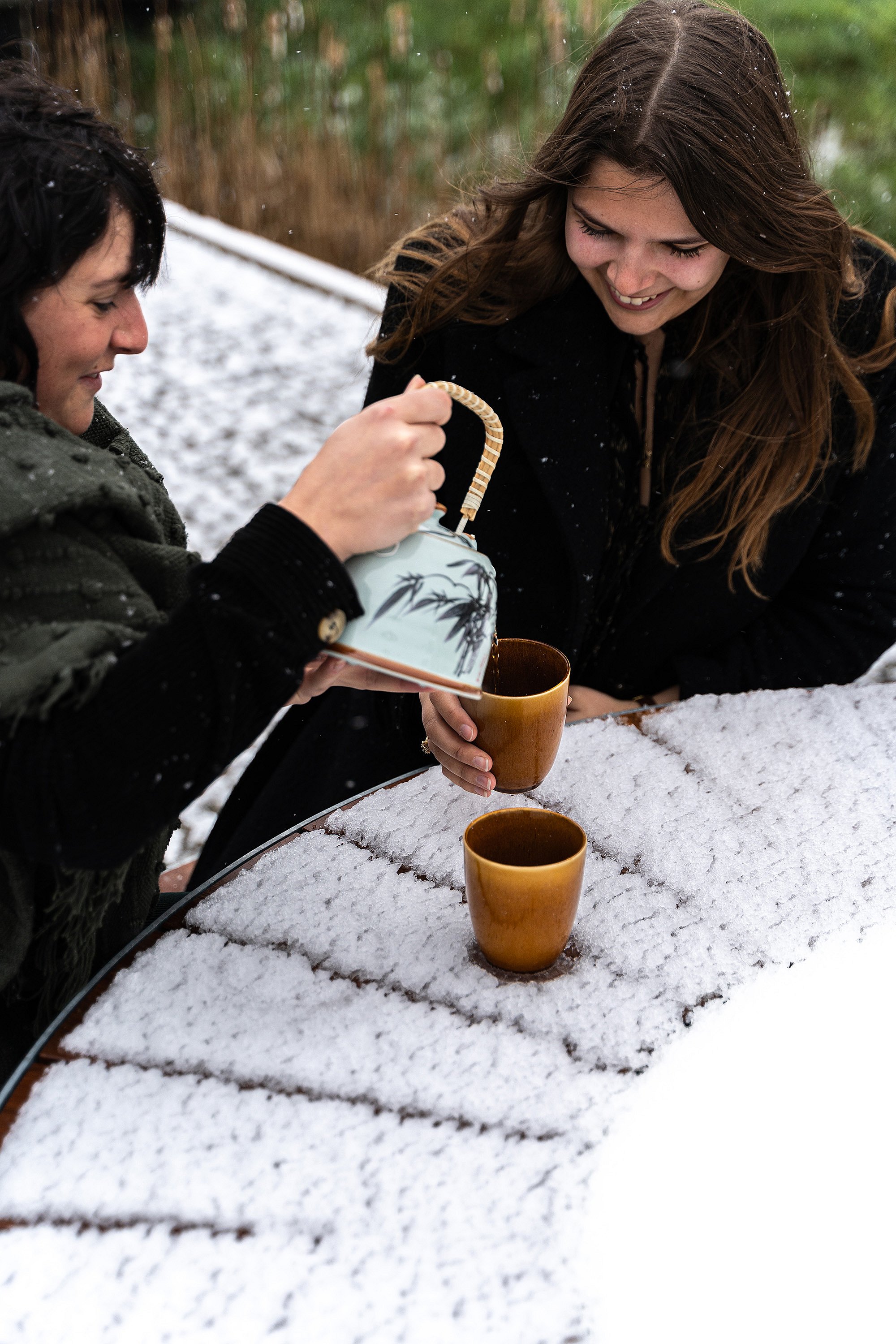 Nahaufnahme von zwei Frauen, die heiße Getränke über die schneebedeckte Holzplatte eines Pantagruel-Picknicktisches gießen und halten.