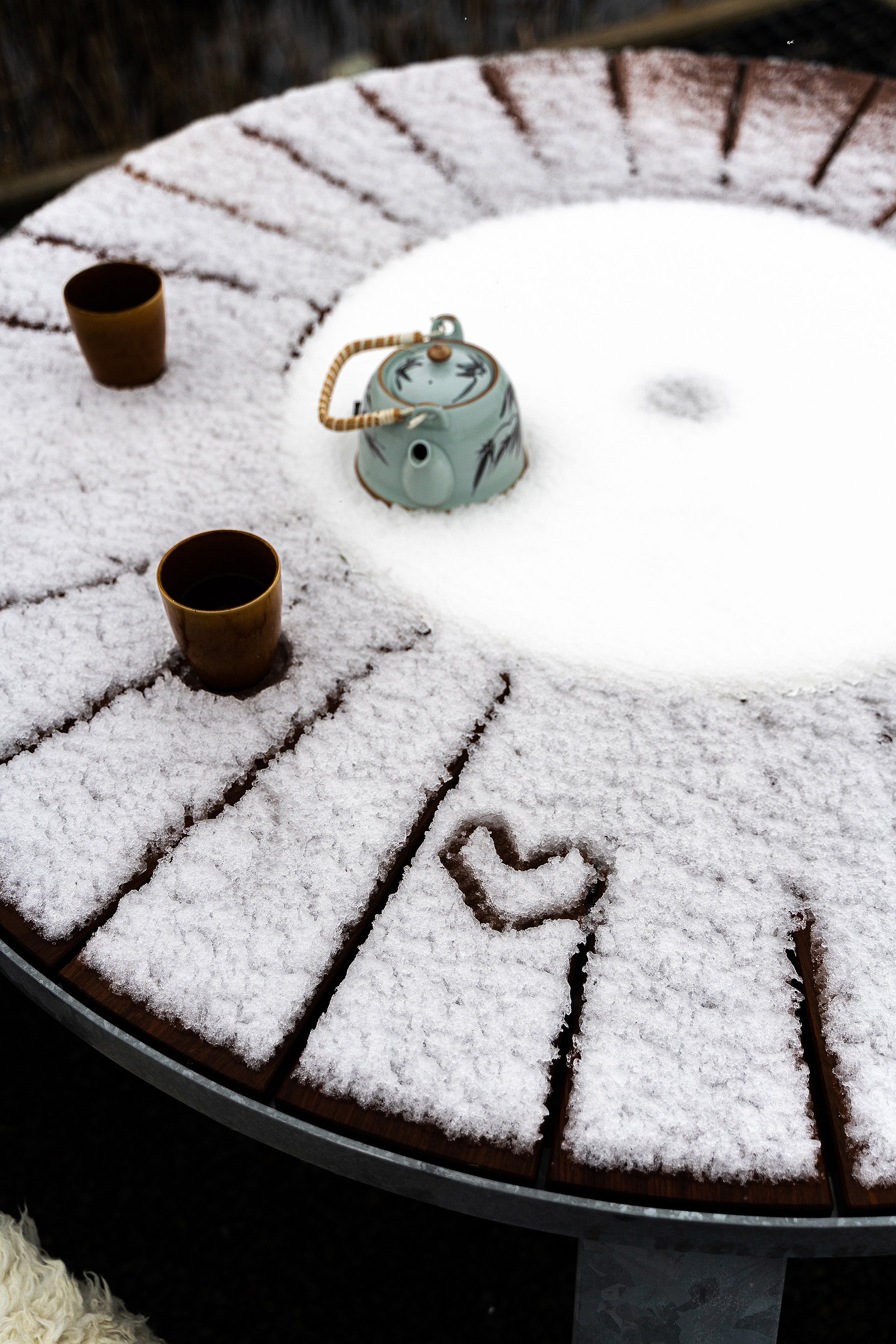 Nahaufnahme einer schneebedeckten Pantagruel-Tischplatte mit zwei Tassen, einer Teekanne und einer kleinen Herzform, die in die verschneite Oberfläche gezeichnet ist.