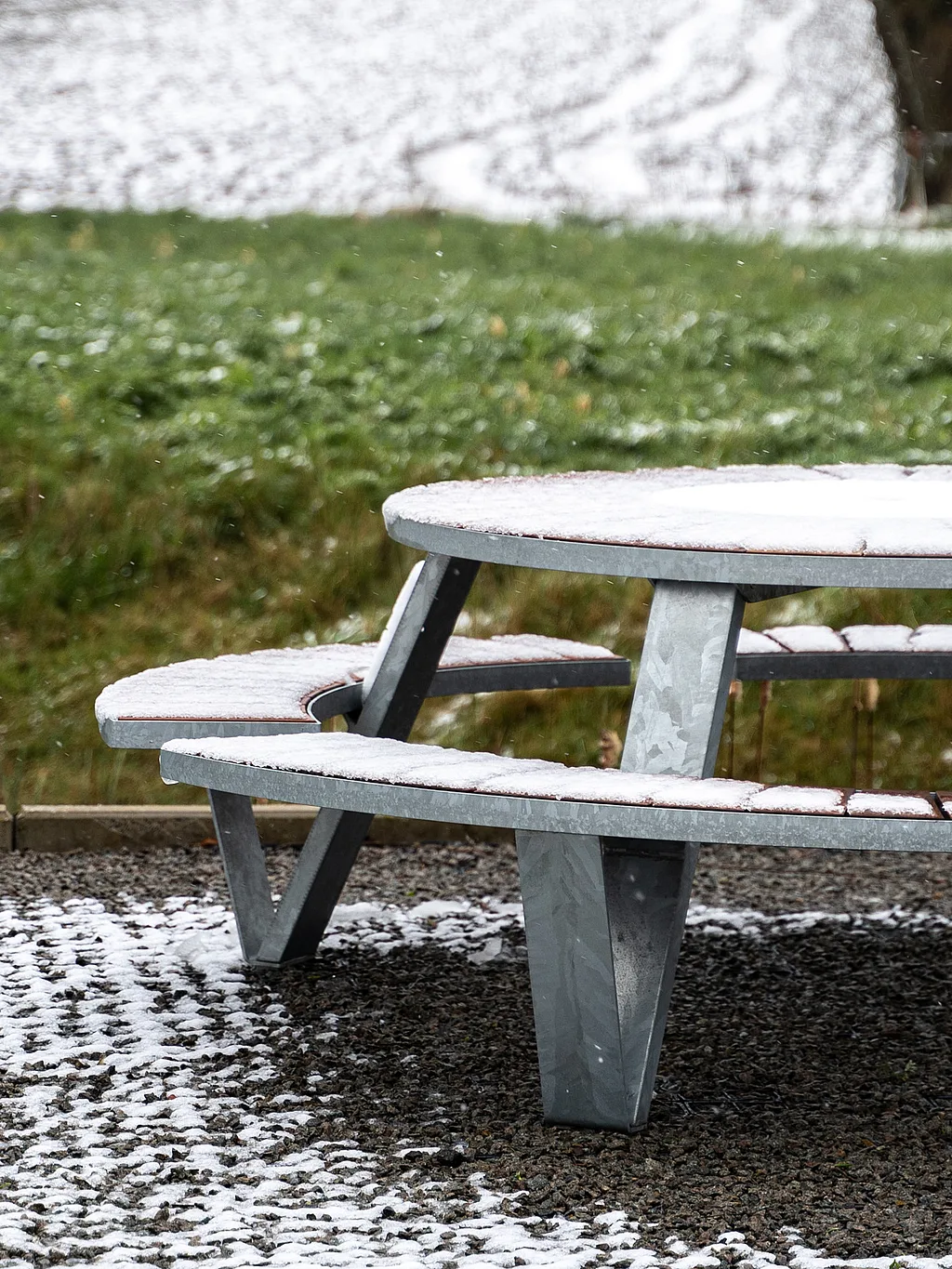 Une table de pique-nique Pantagruel saupoudrée de neige se dresse sur une terrasse de gravier, avec de la neige en petites taches au sol et un paysage herbeux derrière elle.