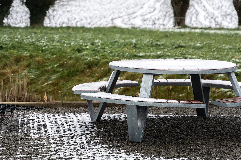 Op een grindterras staat een besneeuwde Pantagruel-picknicktafel, met fragmentarische sneeuw op de grond en een met gras begroeid landschap erachter.