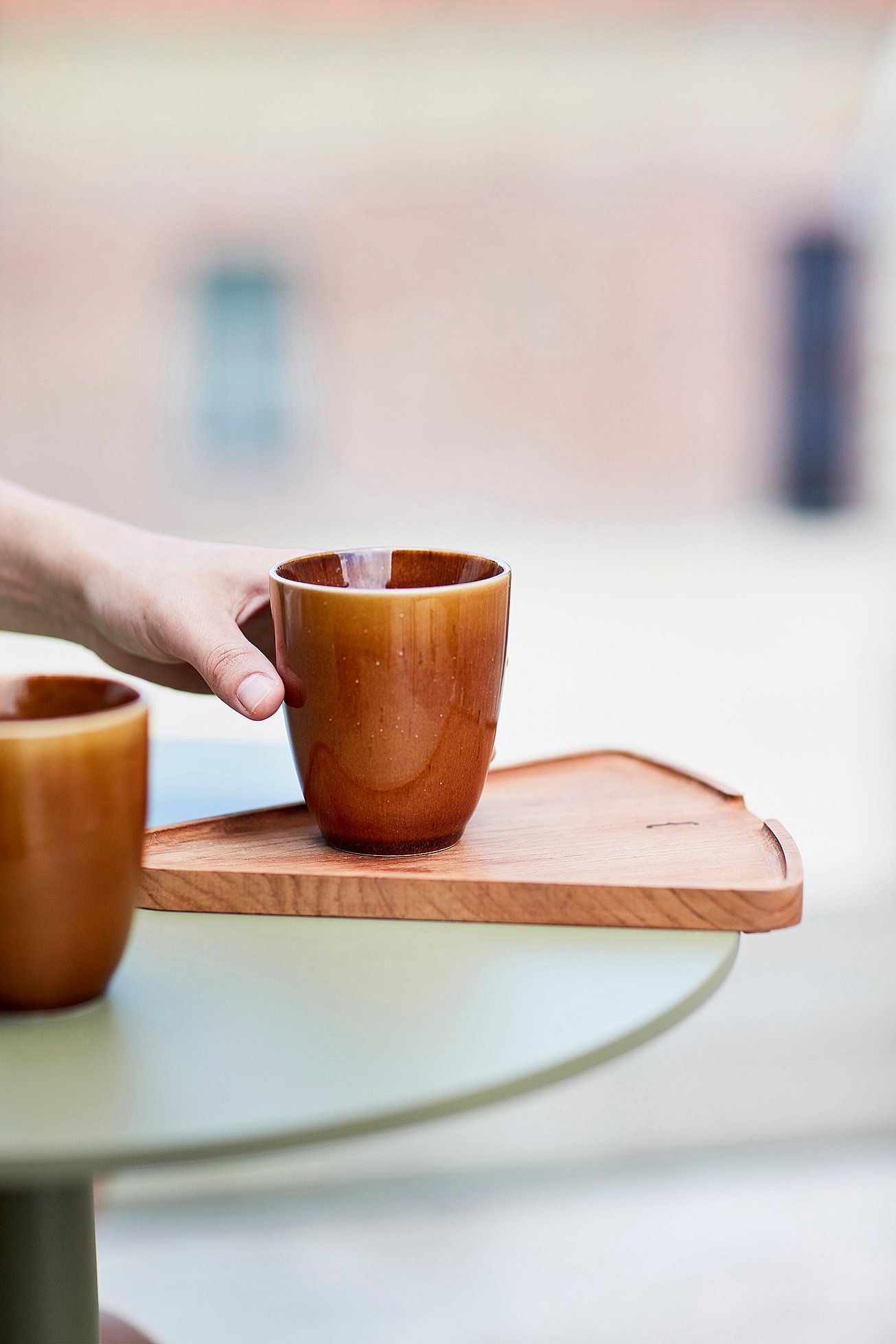   Een hand plaatst een bruine kop op het houten verlengstuk van een rietgroene Bistroo-tafel.