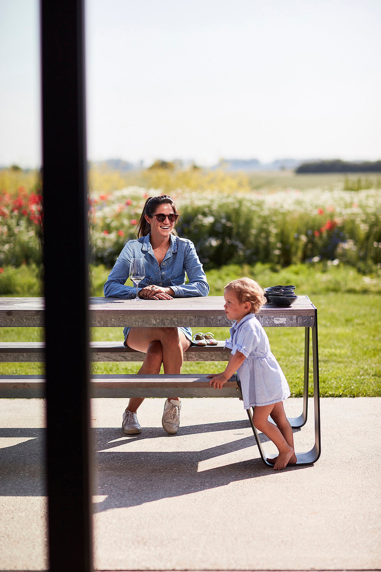   Een vrouw en een kind zitten en leunen tegen een Hopper-picknicktafel op het terras, met wilde bloemen en open landbouwgrond op de achtergrond.