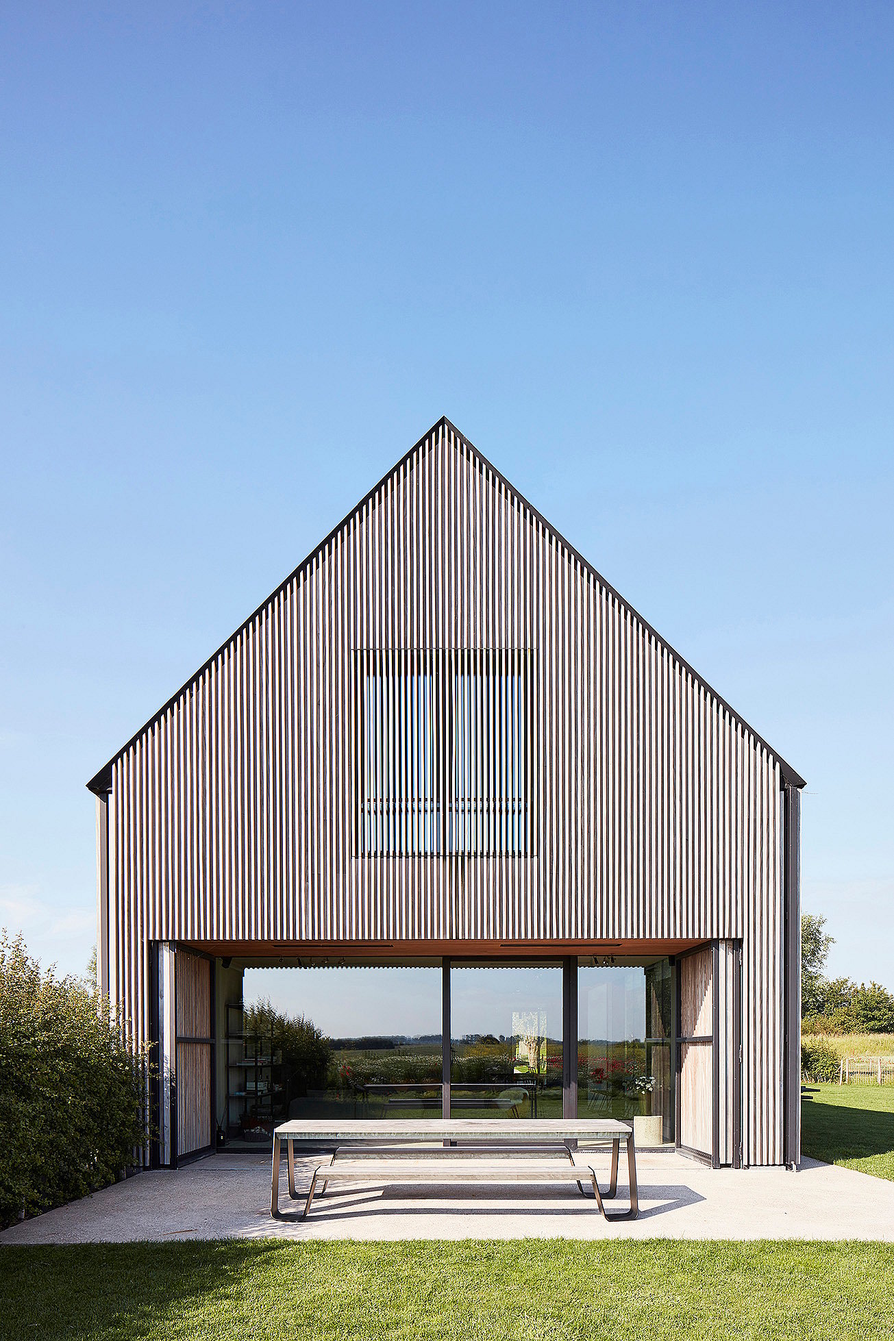 Een met hout bekleed huis met een zadeldak komt uit op een terras met een grote Hopper-picknicktafel voor brede schuifdeuren van glas.