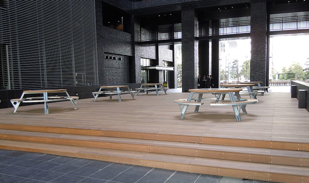 Weite Aussicht auf die große Terrasse im Erdgeschoss von SOHO, wo mehrere Gargantua-Picknicktische auf einem Holzdeck in einem hohen Innenhof aus dunklem Backstein angeordnet sind.