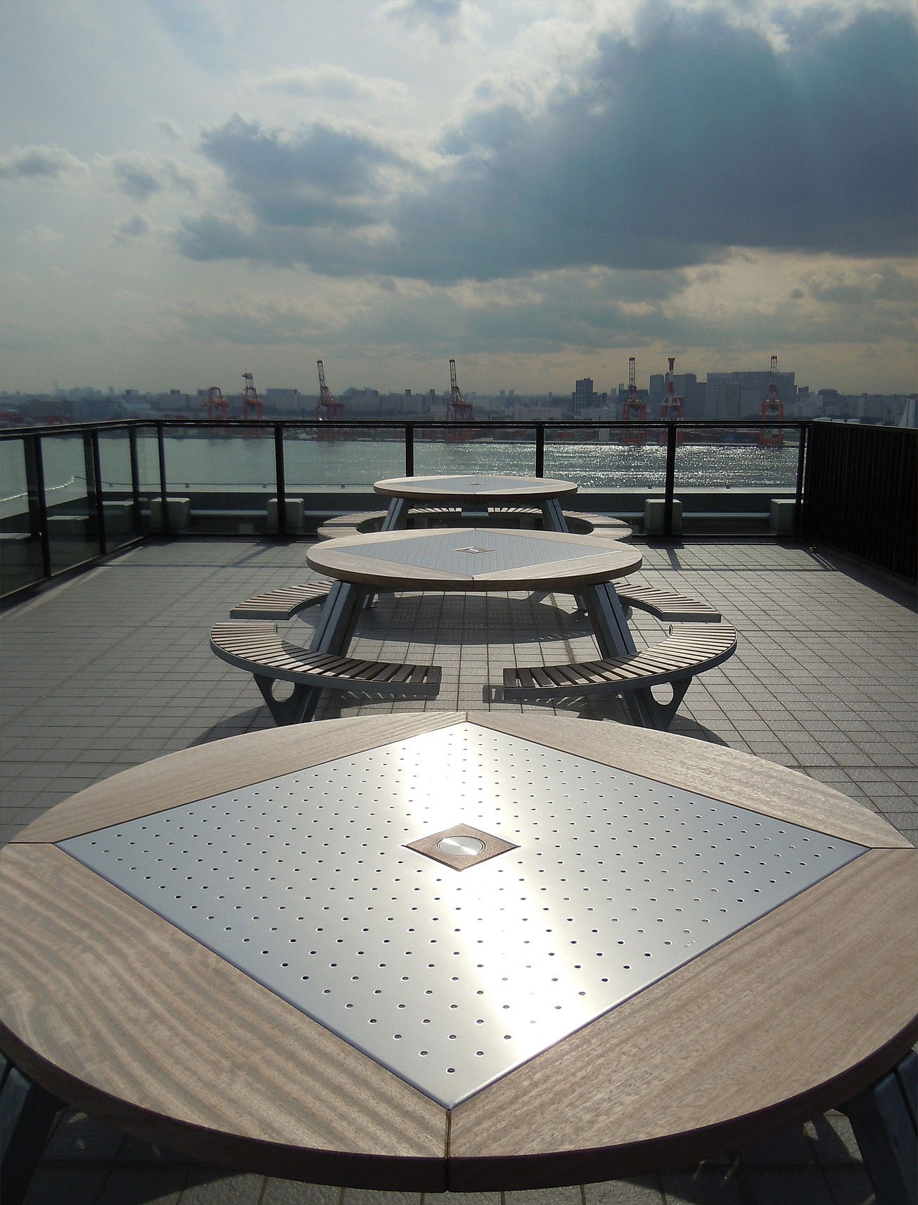 Drei Gargantua-Picknicktische sind auf einer Dachterrasse mit Blick auf das Wasser ausgerichtet, mit Kränen und der Skyline von Tokio in der Ferne.