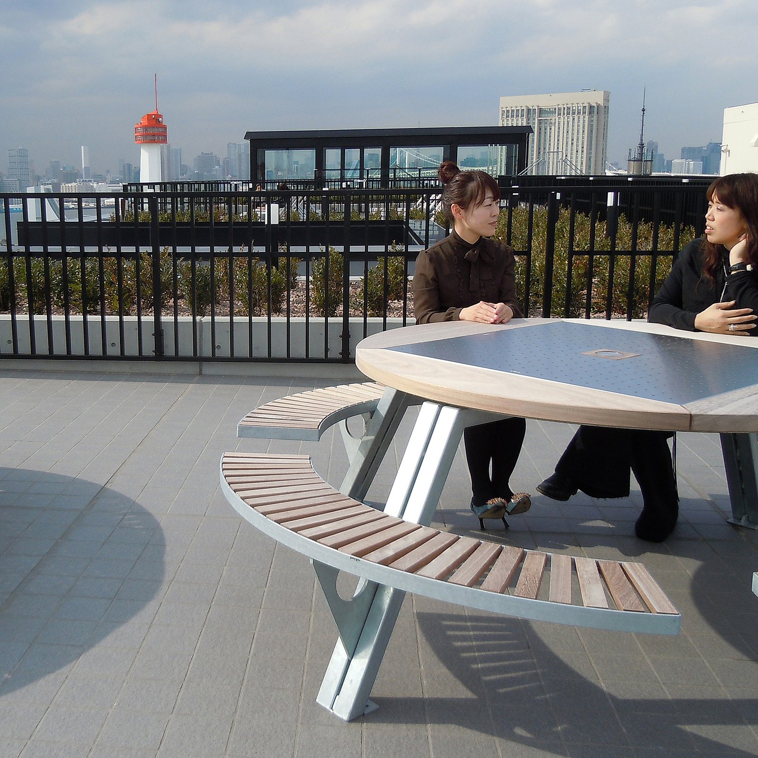 Zwei Frauen sitzen und unterhalten sich an einem Gargantua-Picknicktisch auf der SOHO-Dachterrasse, hinter ihnen die Skyline von Tokio und das schwarze Geländer