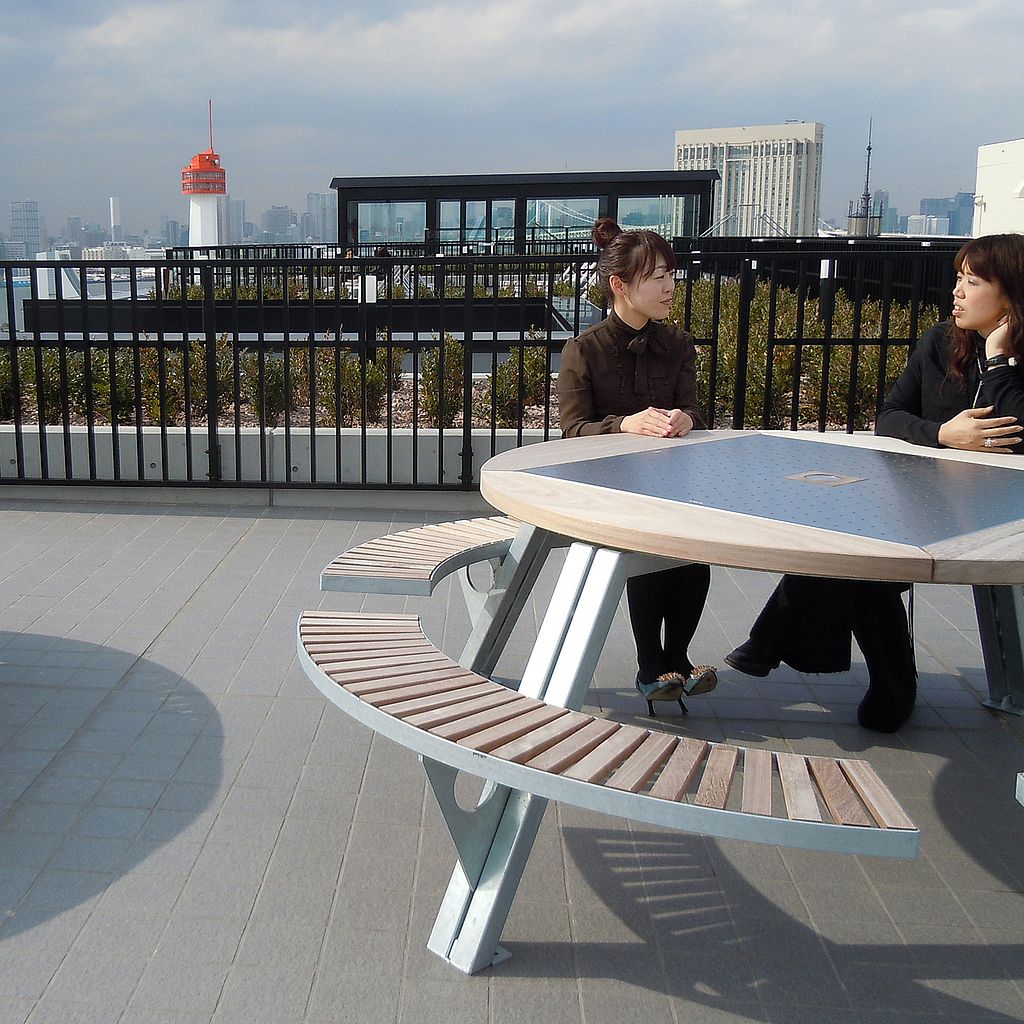 Zwei Frauen sitzen und unterhalten sich an einem Gargantua-Picknicktisch auf der SOHO-Dachterrasse, hinter ihnen die Skyline von Tokio und das schwarze Geländer