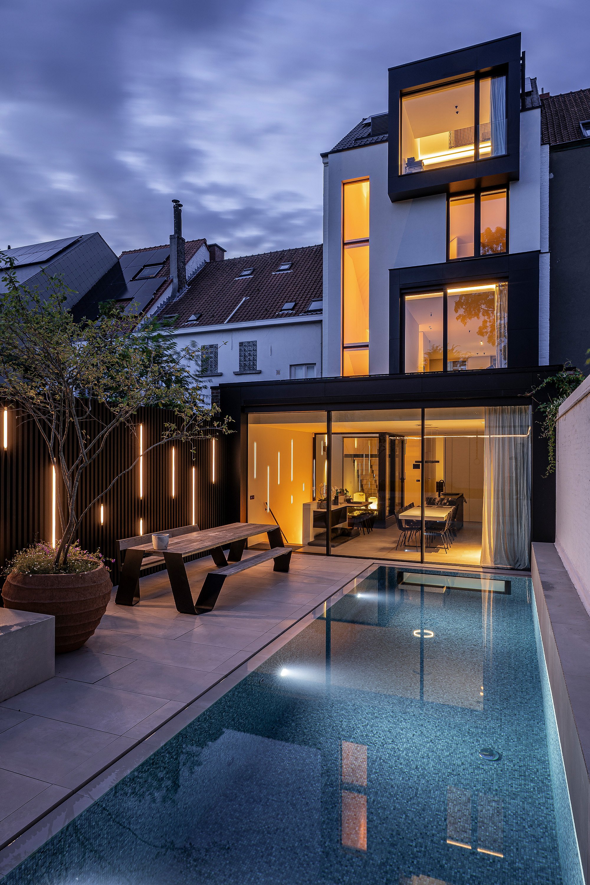 In der Abenddämmerung steht ein Hopper-Picknicktisch neben dem beleuchteten Pool vor der gläsernen Rückfassade des Stadthauses. Warmes Licht aus dem Inneren fällt in den Garten.
