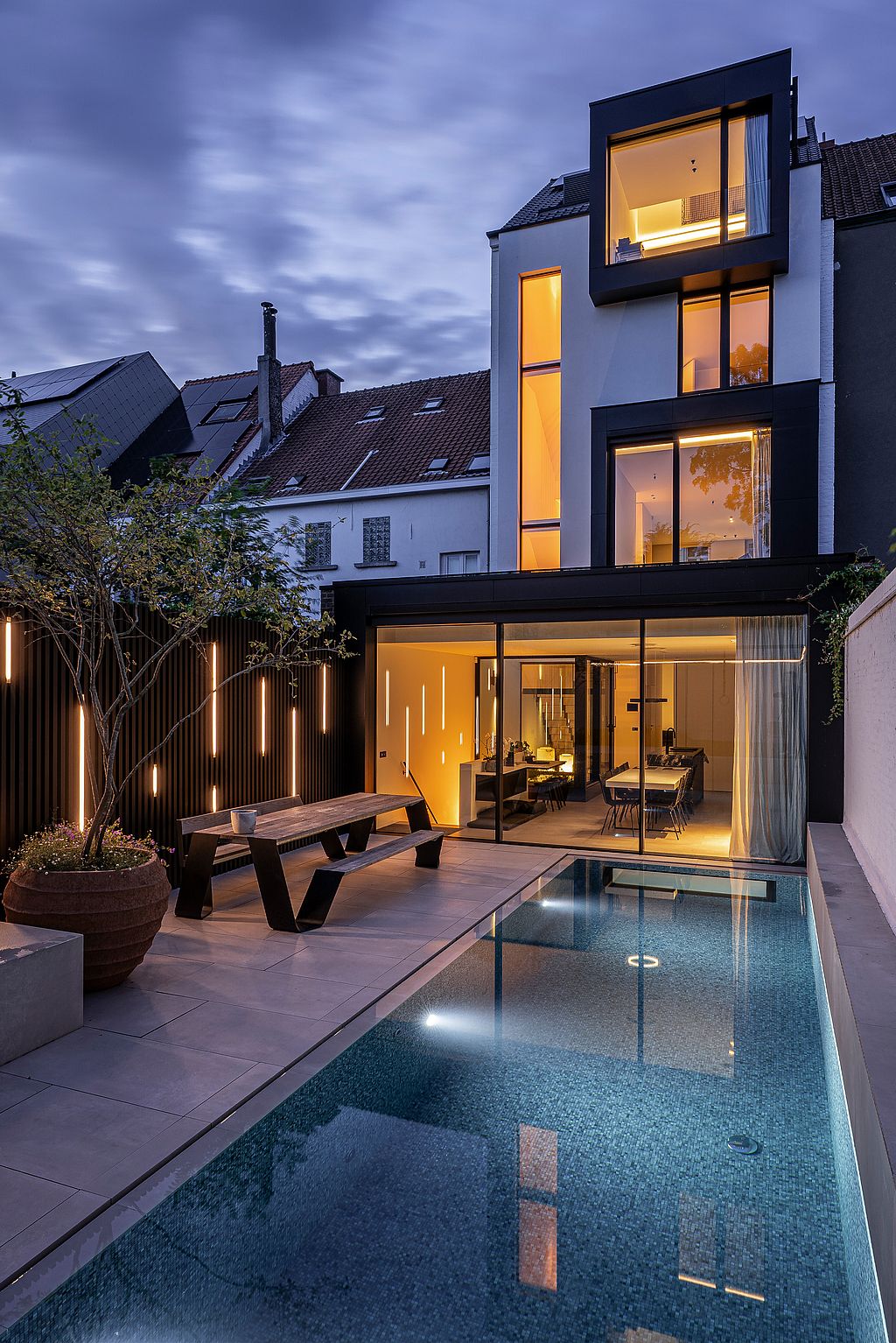In der Abenddämmerung steht ein Hopper-Picknicktisch neben dem beleuchteten Pool vor der gläsernen Rückfassade des Stadthauses. Warmes Licht aus dem Inneren fällt in den Garten.