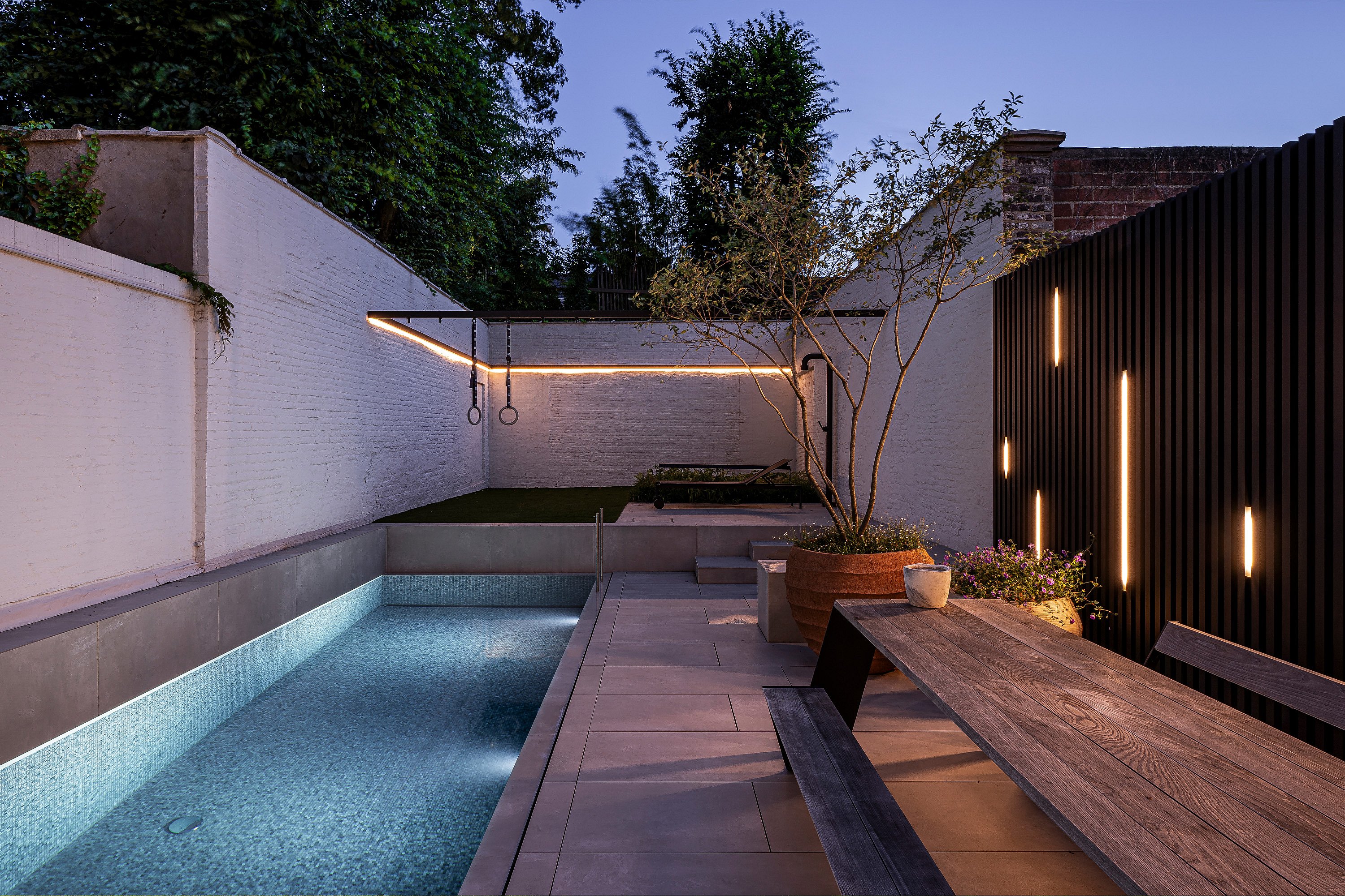 Abends säumt ein Hopper-Picknicktisch den leuchtenden Pool. Am anderen Ende des Gartens befinden sich lineare Wandleuchten, Turnringe und eine schwarze Lounge-Ecke.