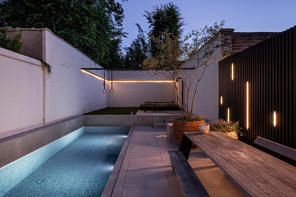 Abends säumt ein Hopper-Picknicktisch den leuchtenden Pool. Am anderen Ende des Gartens befinden sich lineare Wandleuchten, Turnringe und eine schwarze Lounge-Ecke.
