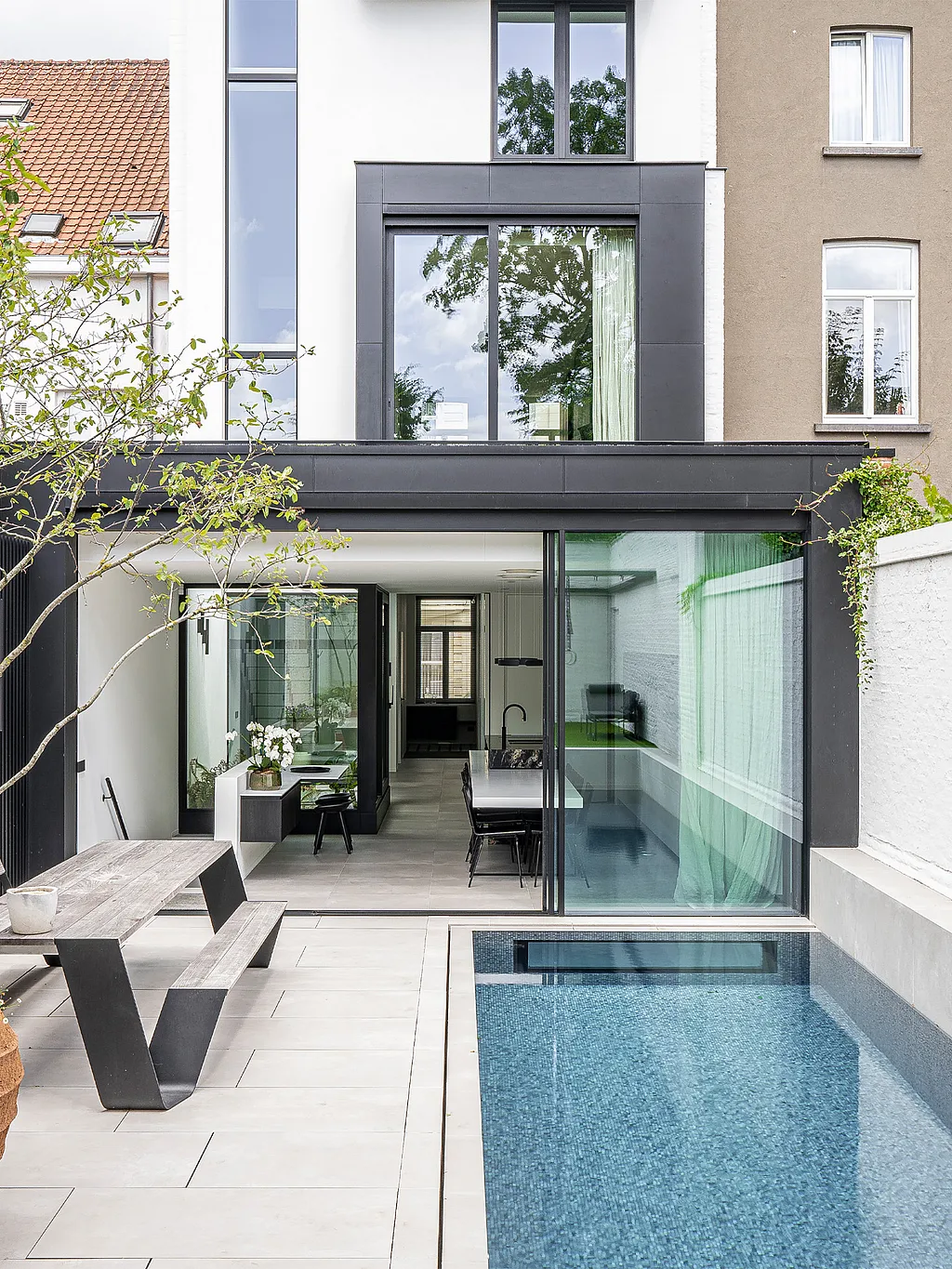 Een smalle stadstuin met een lang zwembad, witte bakstenen muren en een verweerde houten Hopper picknicktafel naast de open achtergevel van een modern herenhuis.