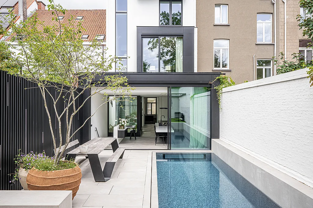 Un jardin urbain étroit avec une longue piscine, des murs de briques blanches et une table de pique-nique Hopper en bois patiné, jouxtant la façade arrière ouverte d'une maison de ville moderne.