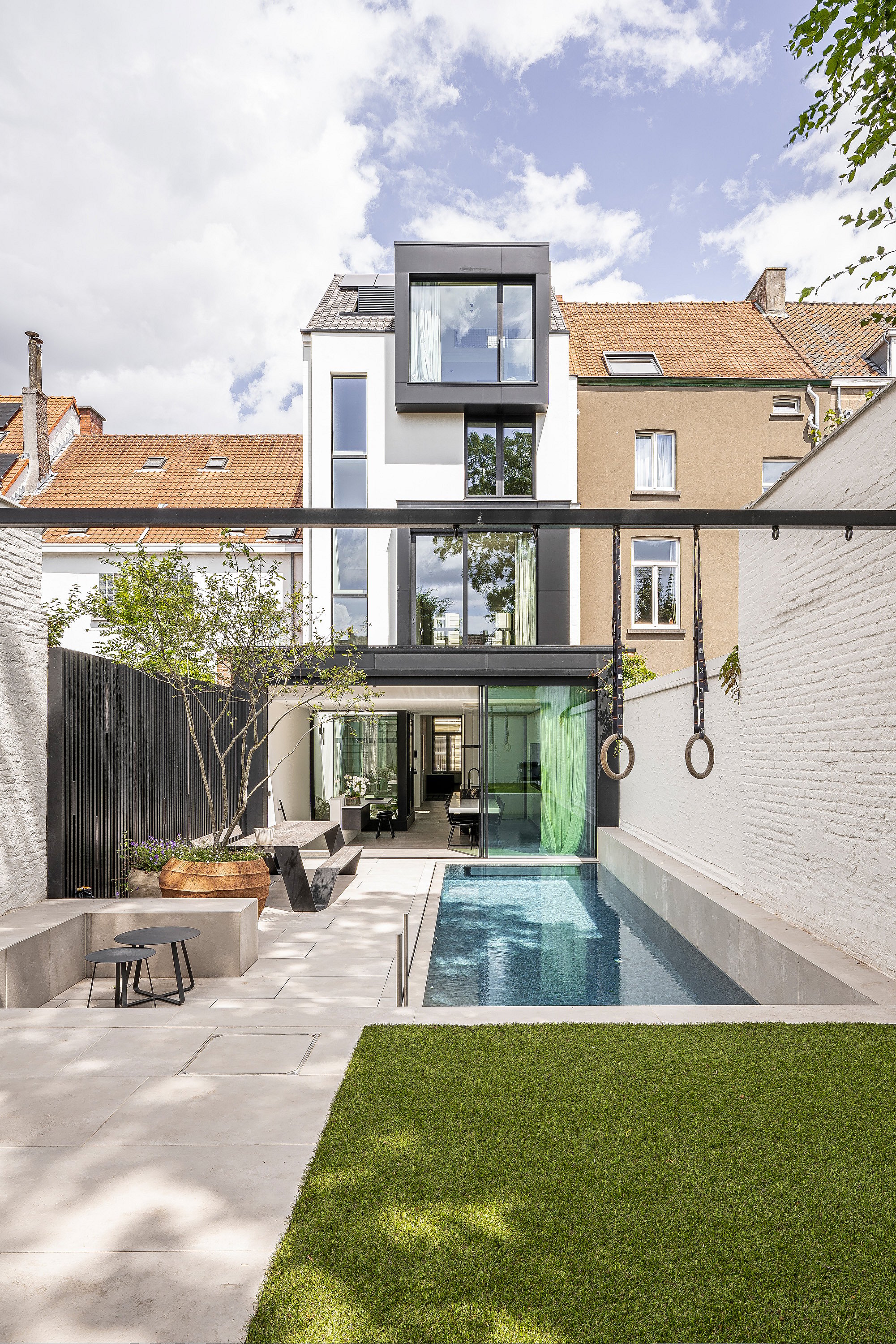   Weitblick auf einen minimalistischen Stadtgarten mit Rasen, Schwimmbecken, Turnringen und einer schwarzen Sol+Luna-Lounge im hinteren Bereich, eingerahmt von hohen weißen Backsteinmauern.