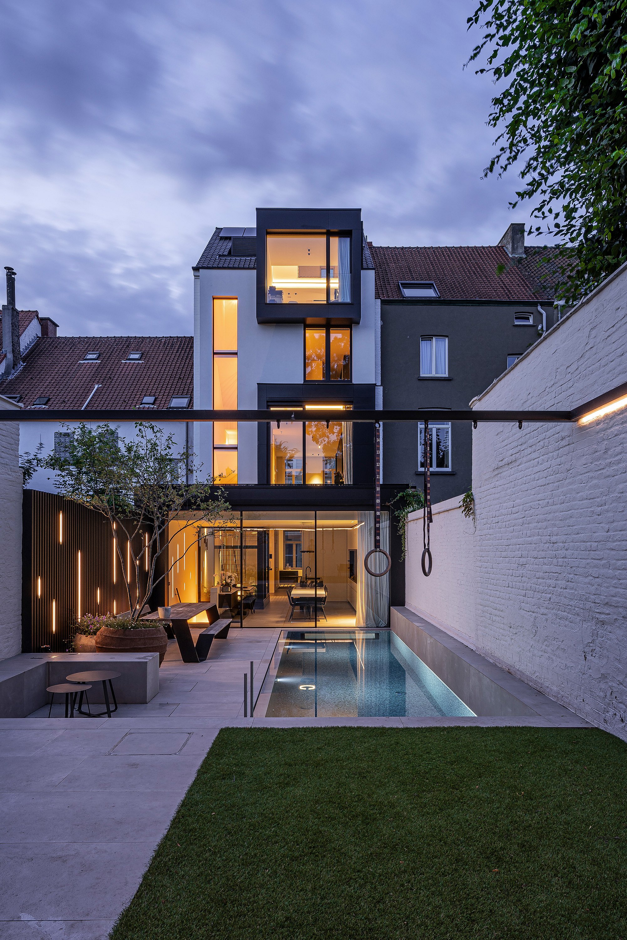 Der Garten des Stadthauses erstrahlt in der Abenddämmerung. Der beleuchtete Pool, die Turnringe und die minimalistische Terrasse bilden eine moderne urbane Oase hinter dem Haus.