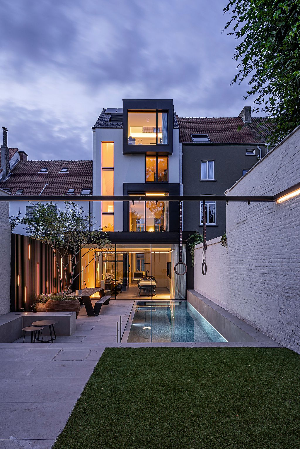 Der Garten des Stadthauses erstrahlt in der Abenddämmerung. Der beleuchtete Pool, die Turnringe und die minimalistische Terrasse bilden eine moderne urbane Oase hinter dem Haus.