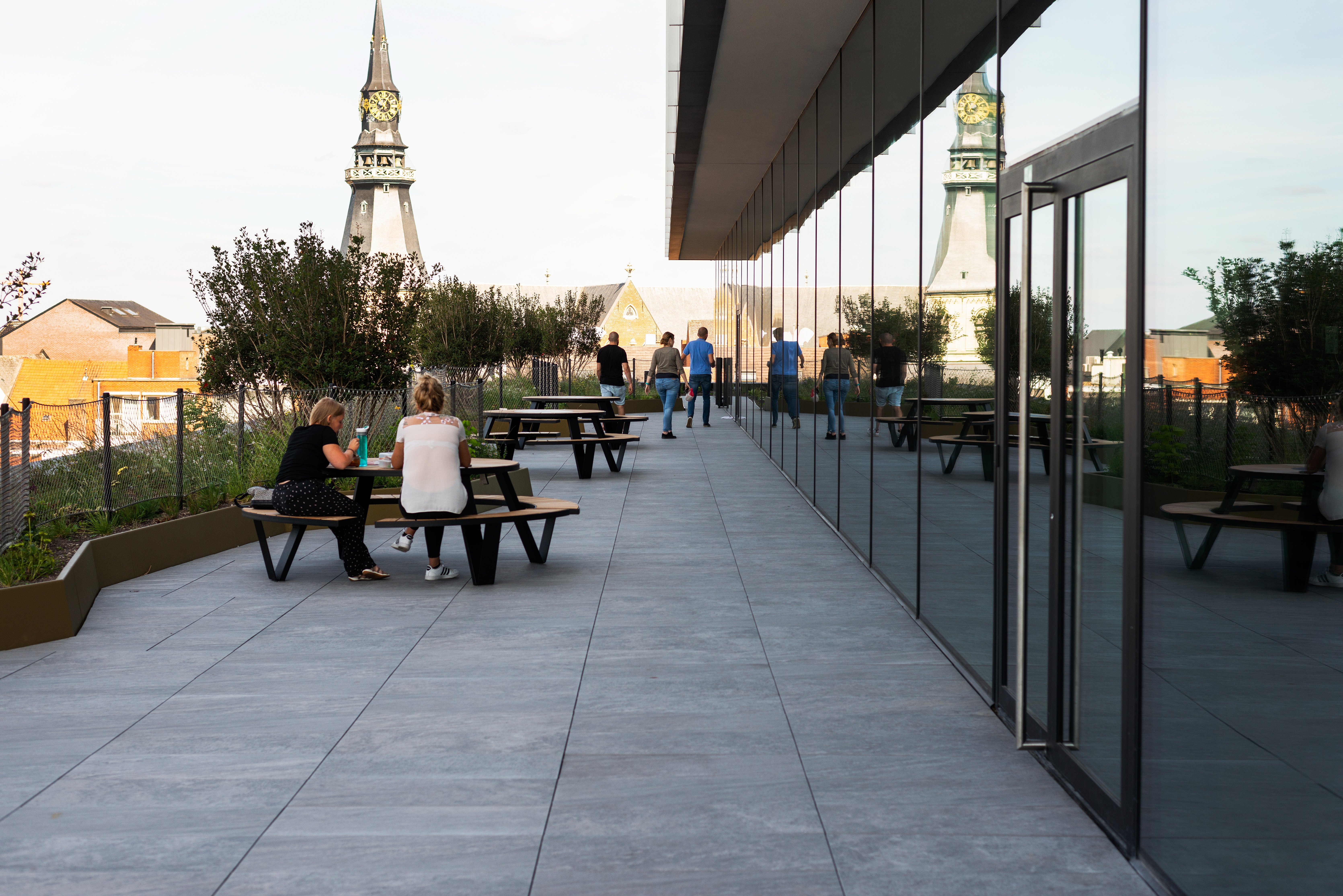 Die Menschen entspannen sich und gehen auf der Dachterrasse neben der reflektierenden Glaswand spazieren. Dahinter sind mehrere Pantagruel-Picknicktische und Kirchtürme zu sehen.