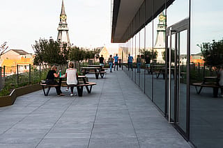 Die Menschen entspannen sich und gehen auf der Dachterrasse neben der reflektierenden Glaswand spazieren. Dahinter sind mehrere Pantagruel-Picknicktische und Kirchtürme zu sehen.
