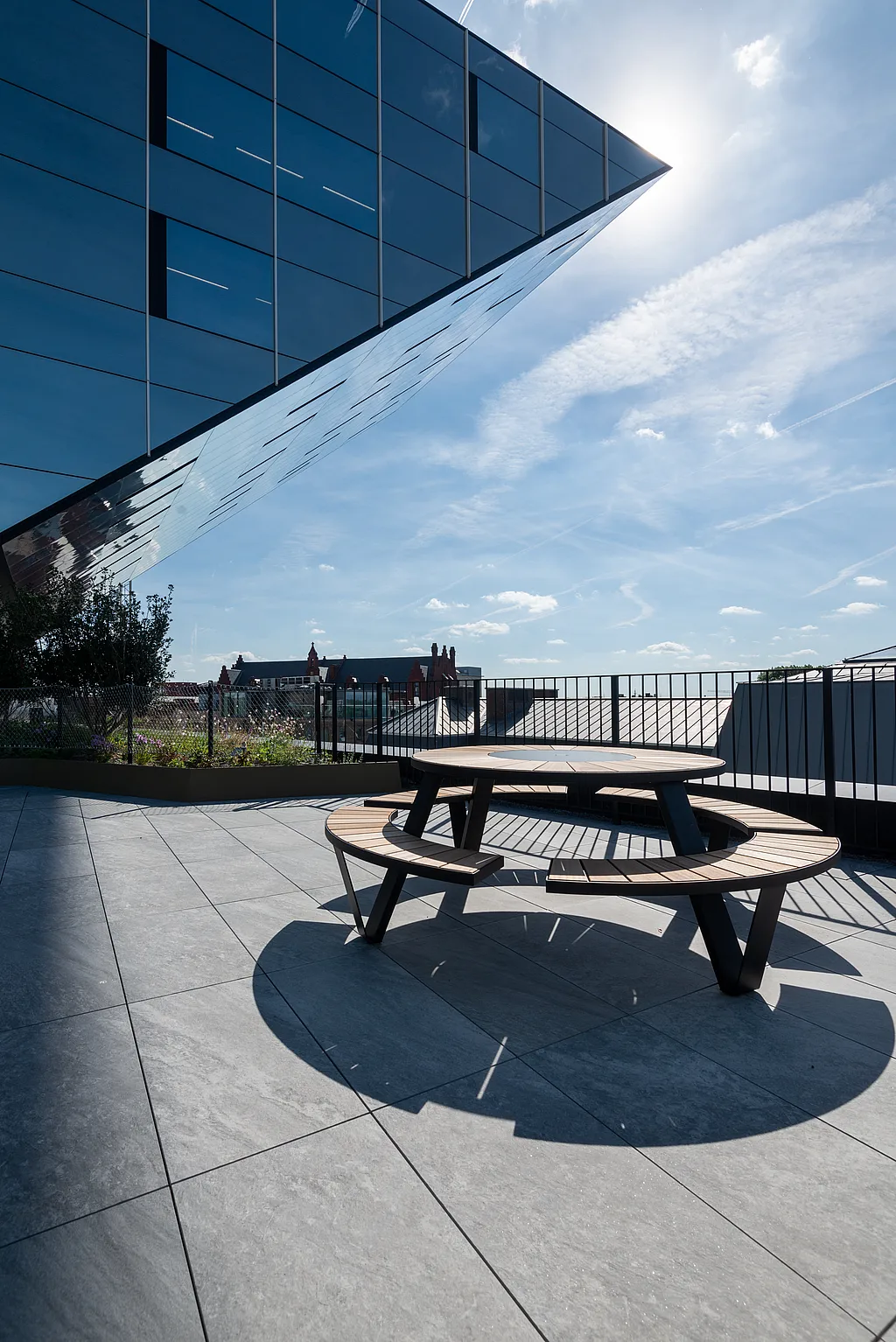 Auf der Dachterrasse unter dem dramatischen Spiegelüberhang des Rathauses von Hasselt steht ein Picknicktisch von Pantagruel, im Hintergrund sind die Skyline und die Bepflanzung zu sehen.