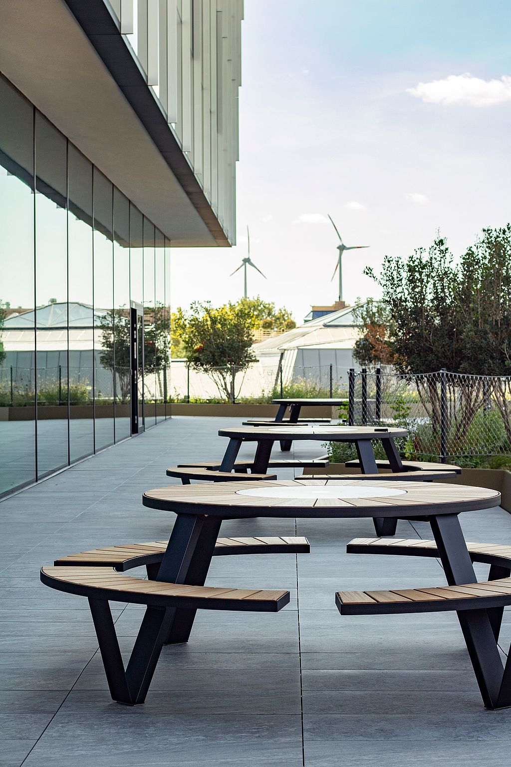 Eine Reihe von Pantagruel-Picknicktischen säumt die Terrasse neben der Glasfassade, in der Ferne Bäume, Bepflanzungen und Windkraftanlagen.