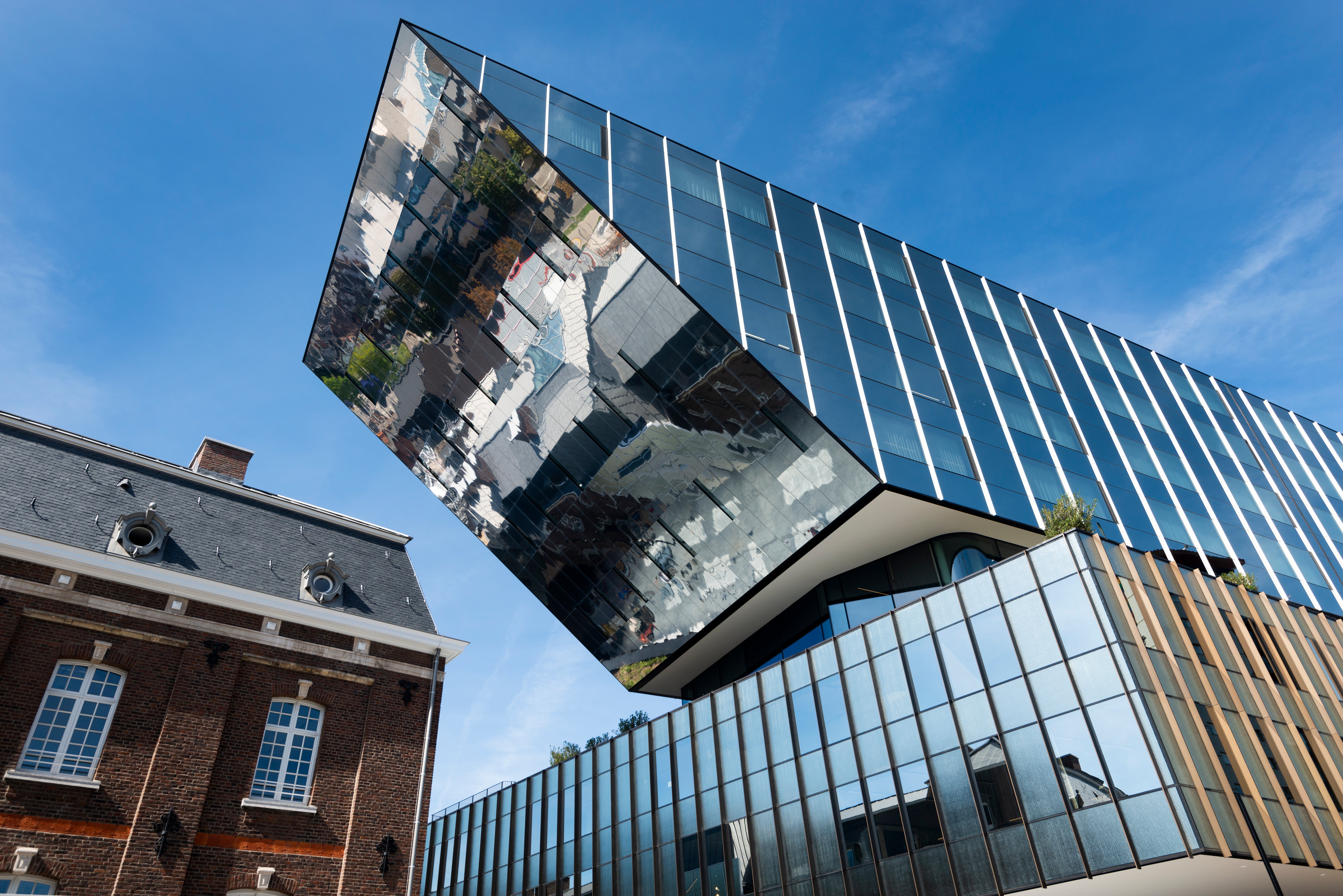 Flachwinkelansicht der verspiegelten Rathausfassade von Hasselt, die das historische Backsteingebäude daneben vor einem klaren blauen Himmel widerspiegelt.