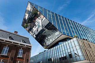 Flachwinkelansicht der verspiegelten Rathausfassade von Hasselt, die das historische Backsteingebäude daneben vor einem klaren blauen Himmel widerspiegelt.