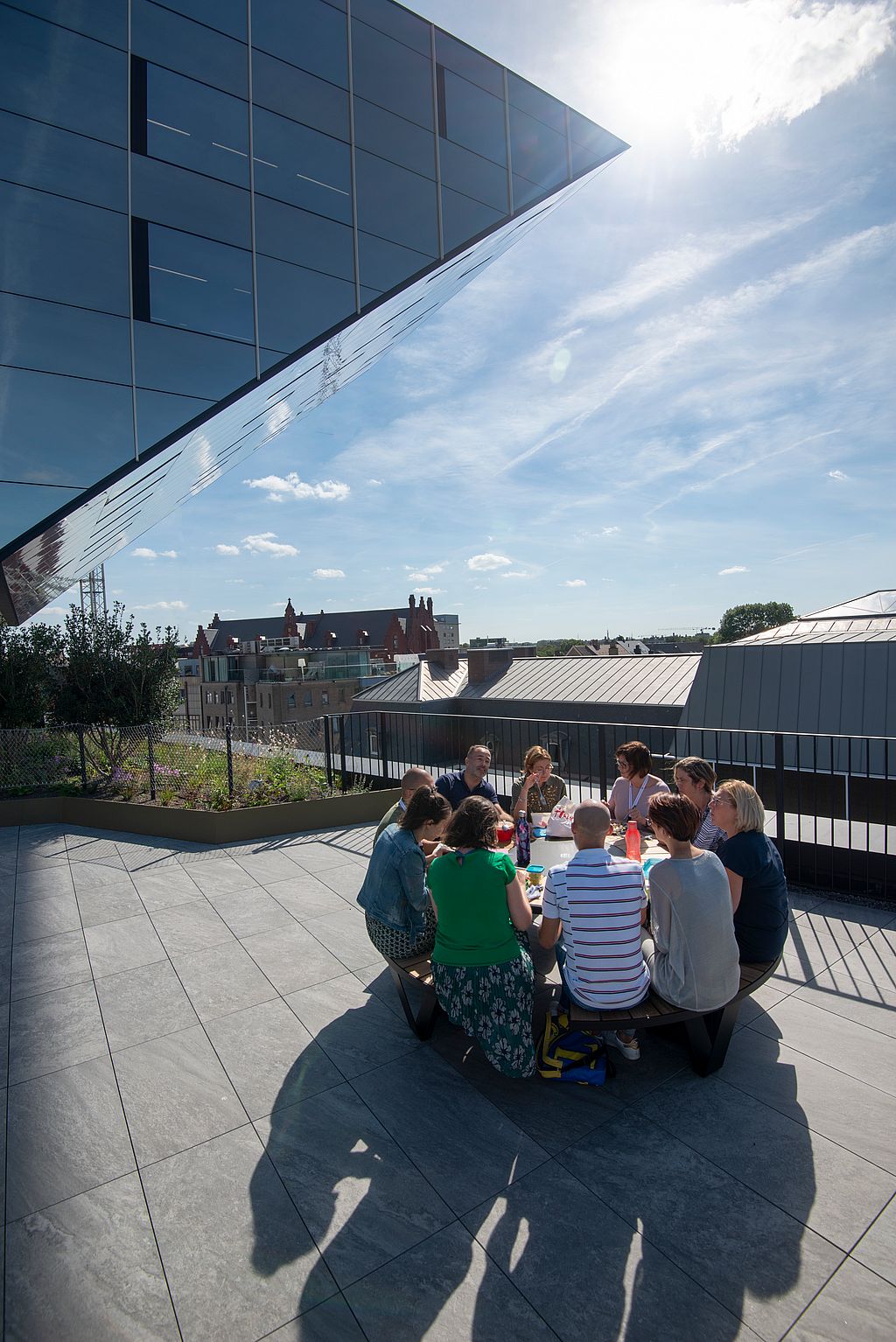 Eine Gruppe versammelt sich um einen Pantagruel-Picknicktisch auf der Dachterrasse und sitzt unter der scharfen Spiegelkante des Rathauses.