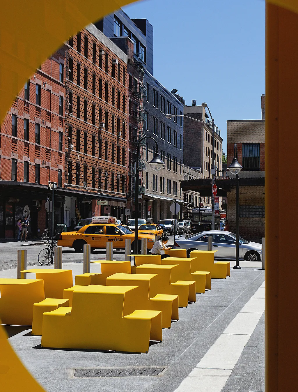 Reihen leuchtend gelber Picnik-Tische säumen den Bürgersteig vor dem Standard Hotel in New York, im Hintergrund ein gelbes Taxi und Backsteingebäude der Stadt.