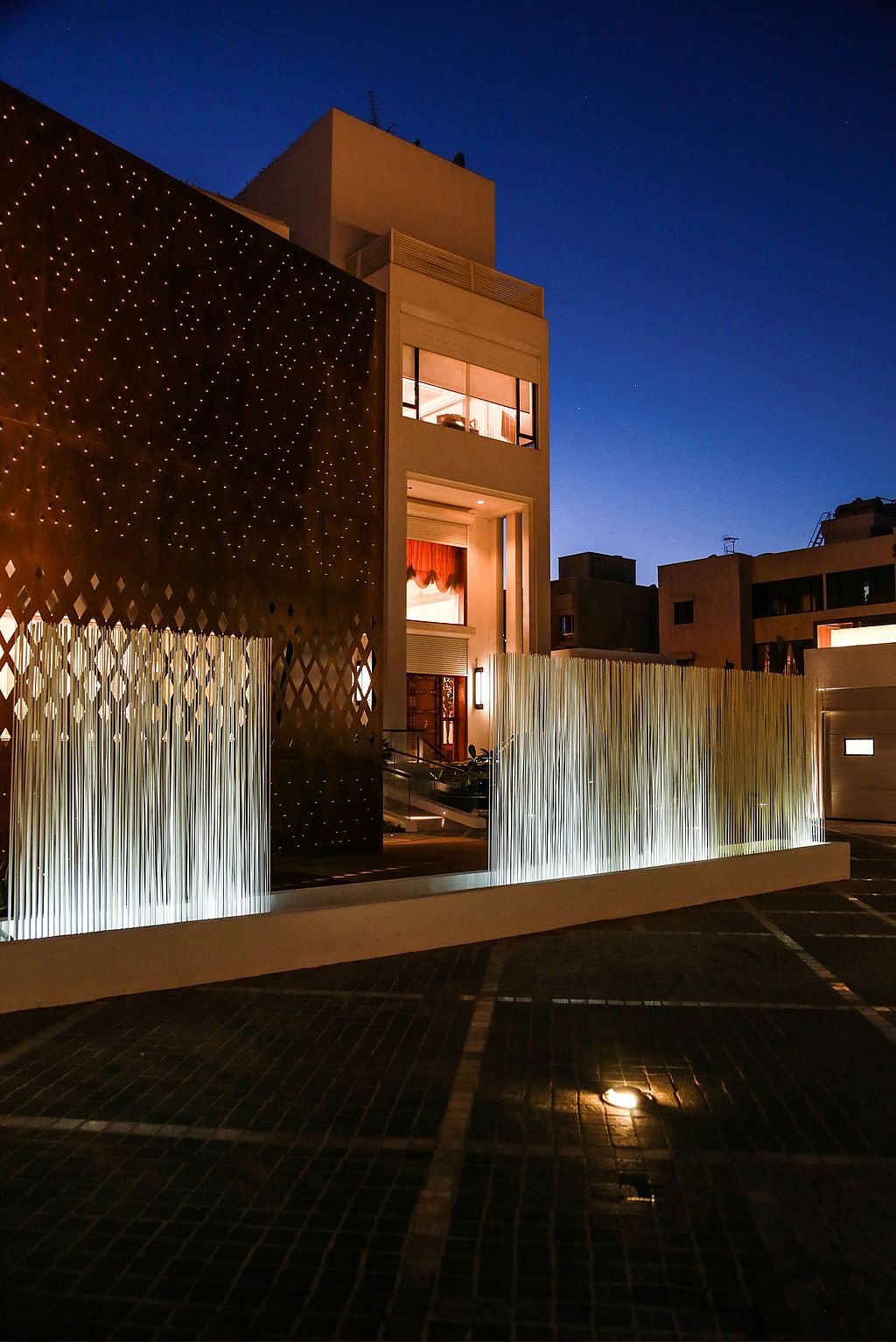 Night view of the diwaniya entrance, with glowing white Sticks framing the driveway in front of the rust-coloured façade and tall white house.