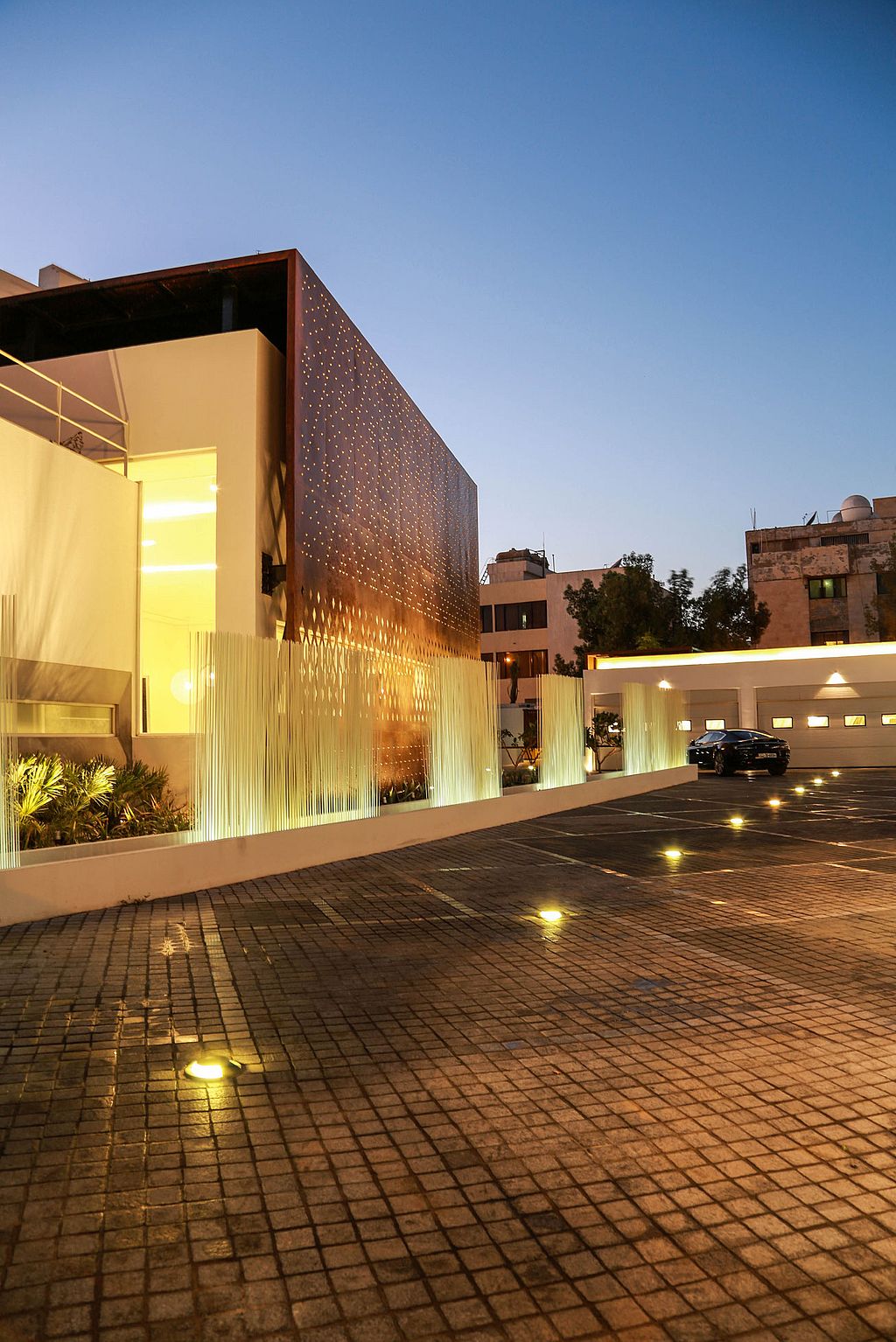 At blue hour, white Sticks stretch along the edge of the driveway beside the illuminated façade, with recessed lights reflected in the paving.
