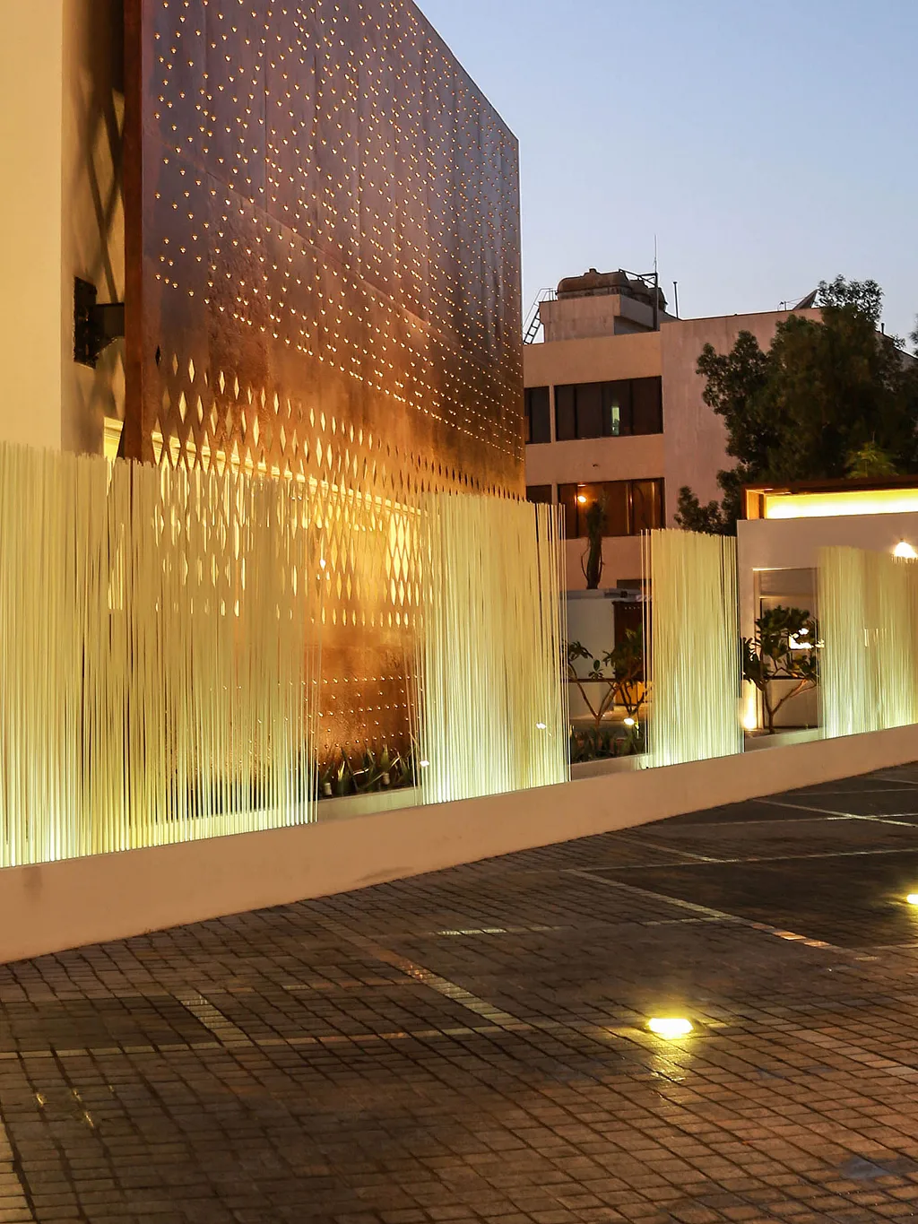 White Sticks light installations line the driveway of a modern Kuwaiti diwaniya at dusk, set against a rust-coloured perforated steel façade and a broad paved forecourt.
