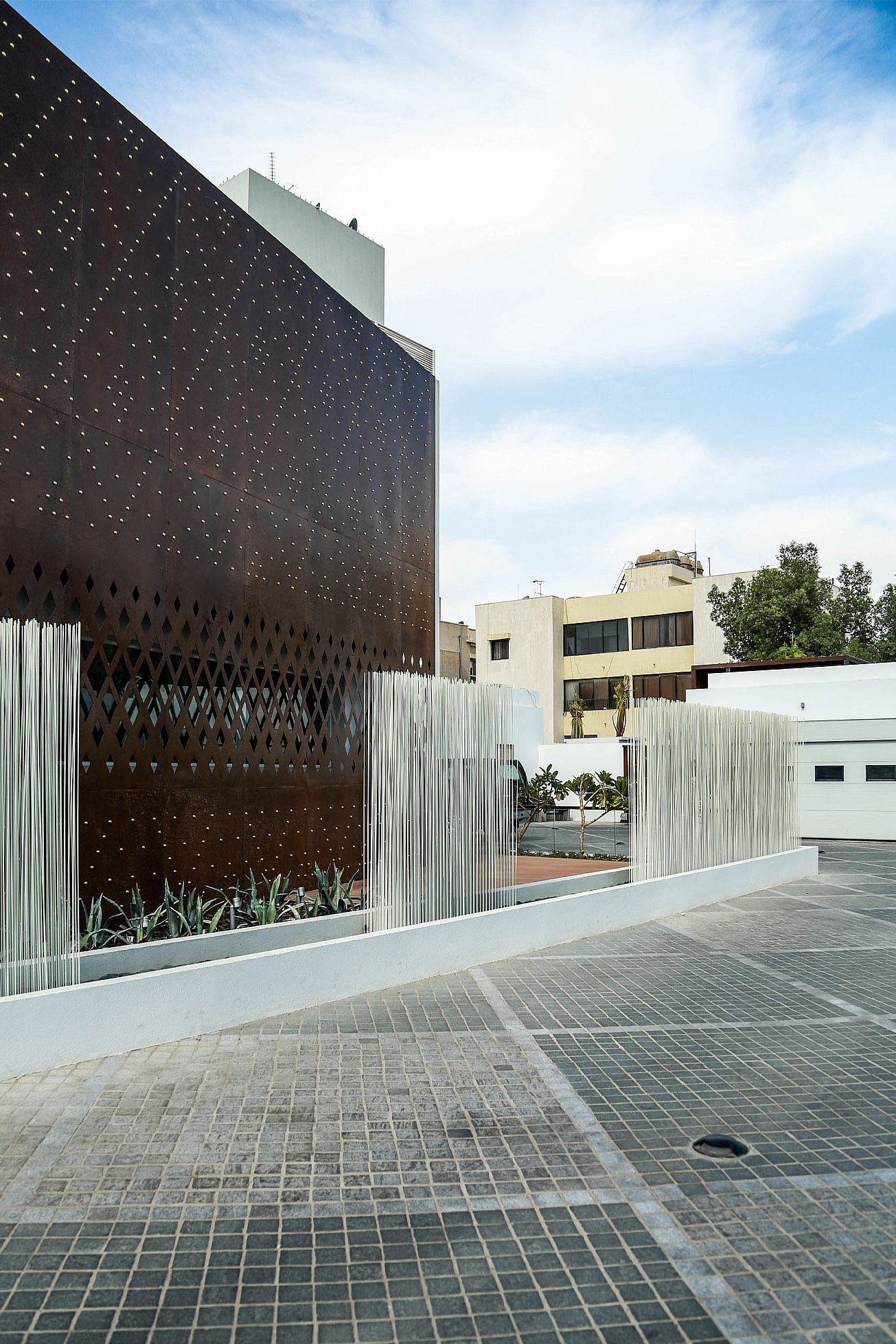 White Sticks rise in separate clusters along the driveway beside the rust-coloured façade, with neighbouring buildings and an open sky in the background.